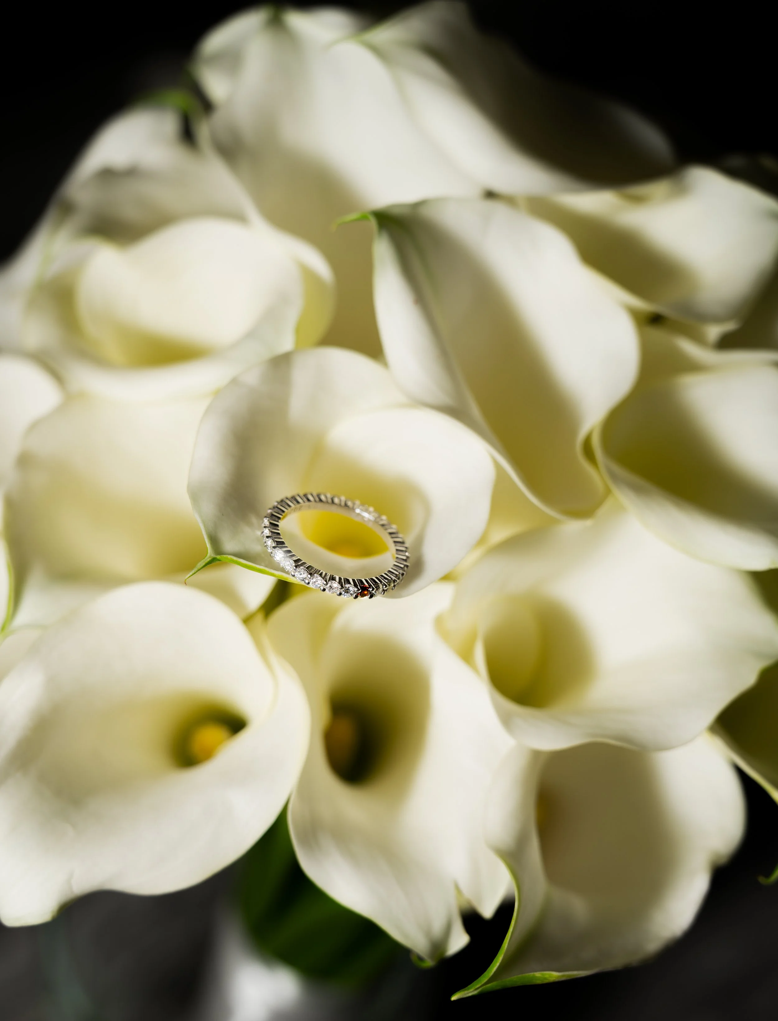 A wedding ring with diamonds placed on white calla lily flowers with soft lighting.
