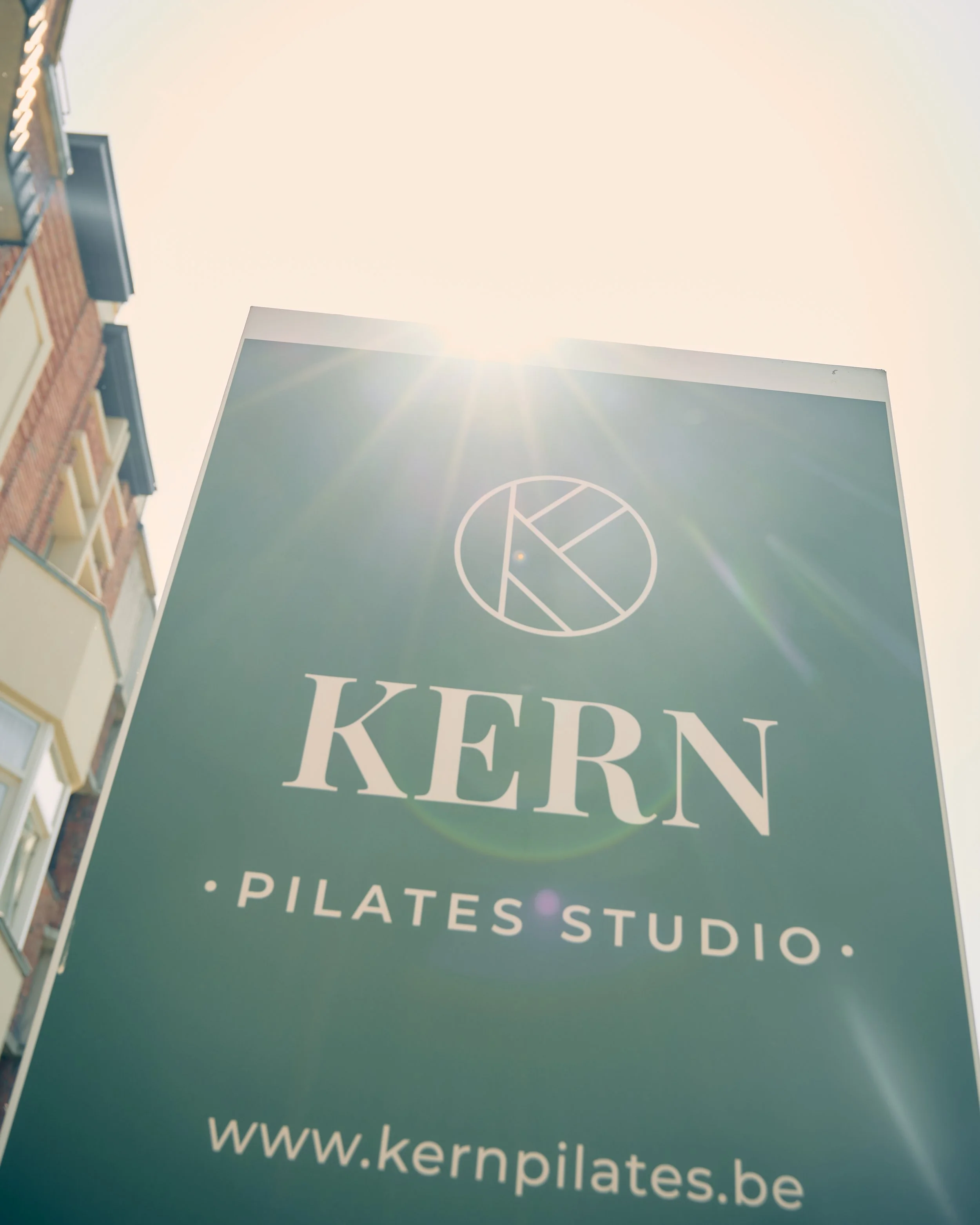 A large teal sign with the words 'KERN' and 'PILATES STUDIO' along with the website 'www.kernpilates.be'. The sun is shining brightly from above, creating lens flare in the image. Part of a brick building with windows is visible on the left.