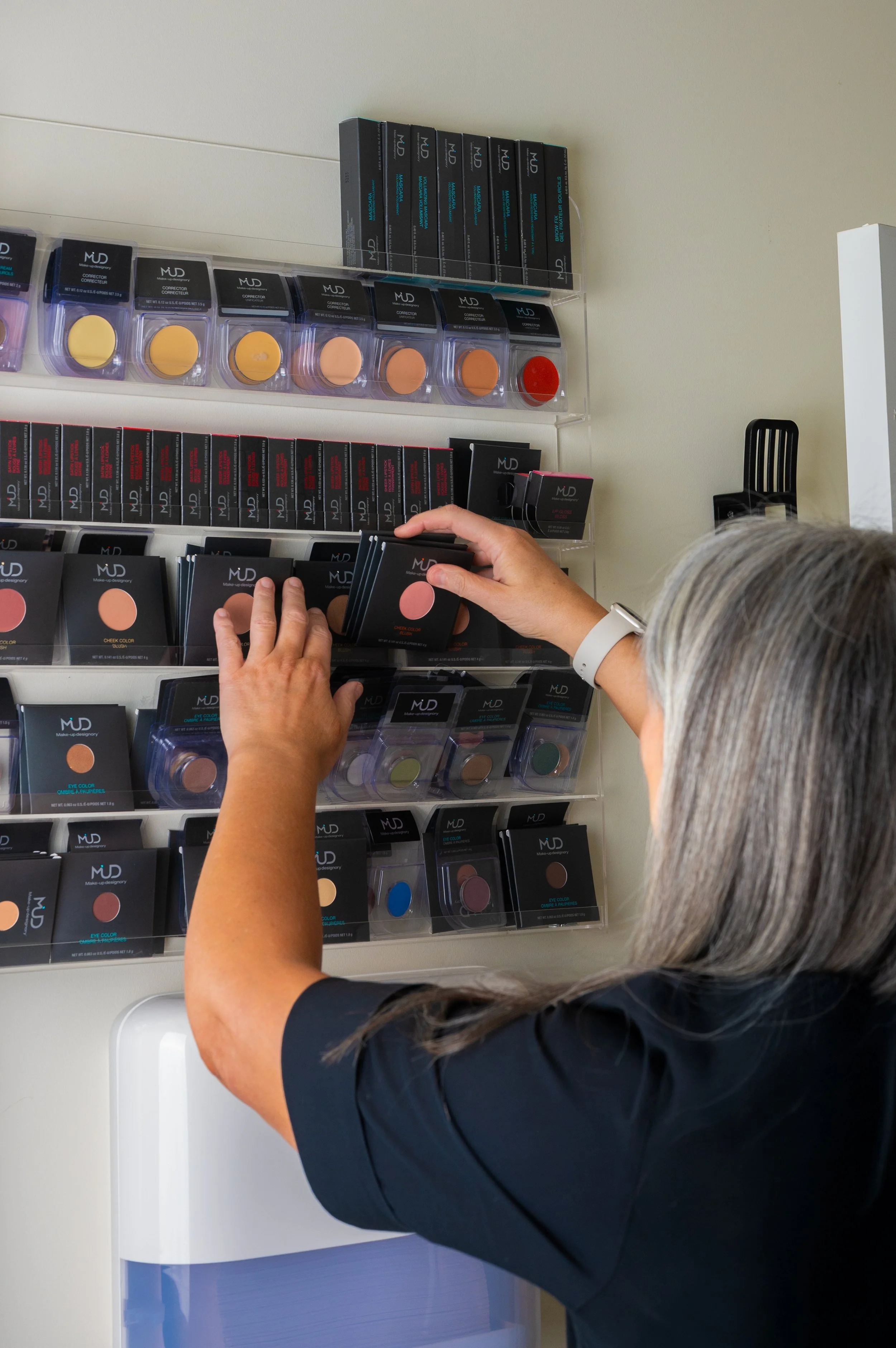 A person with gray hair selecting makeup blush from a display shelf, which includes various shades of powders and other makeup products.