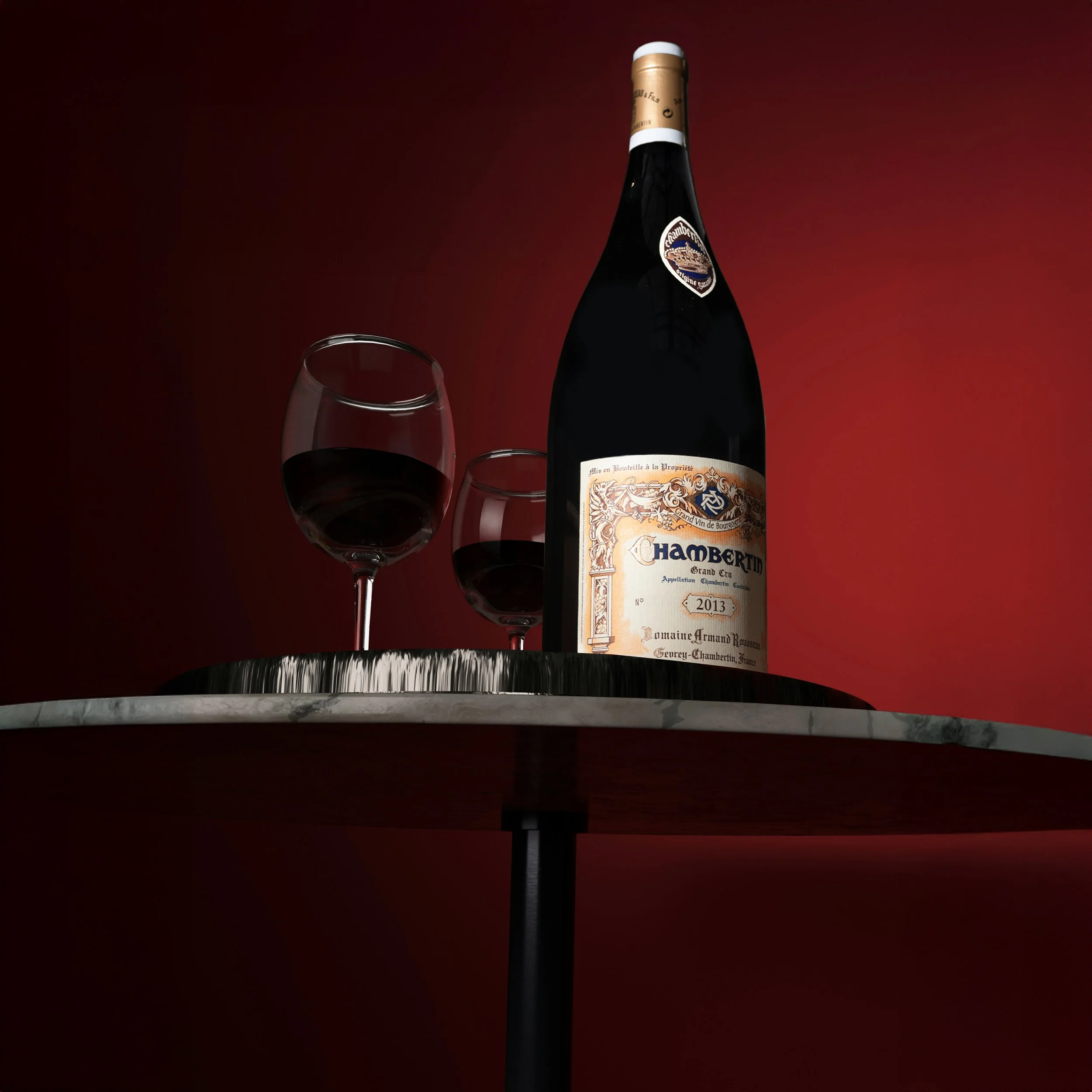 A bottle of Hambert crimson wine with two glasses filled with red wine on a marble table, against a dark red background.
