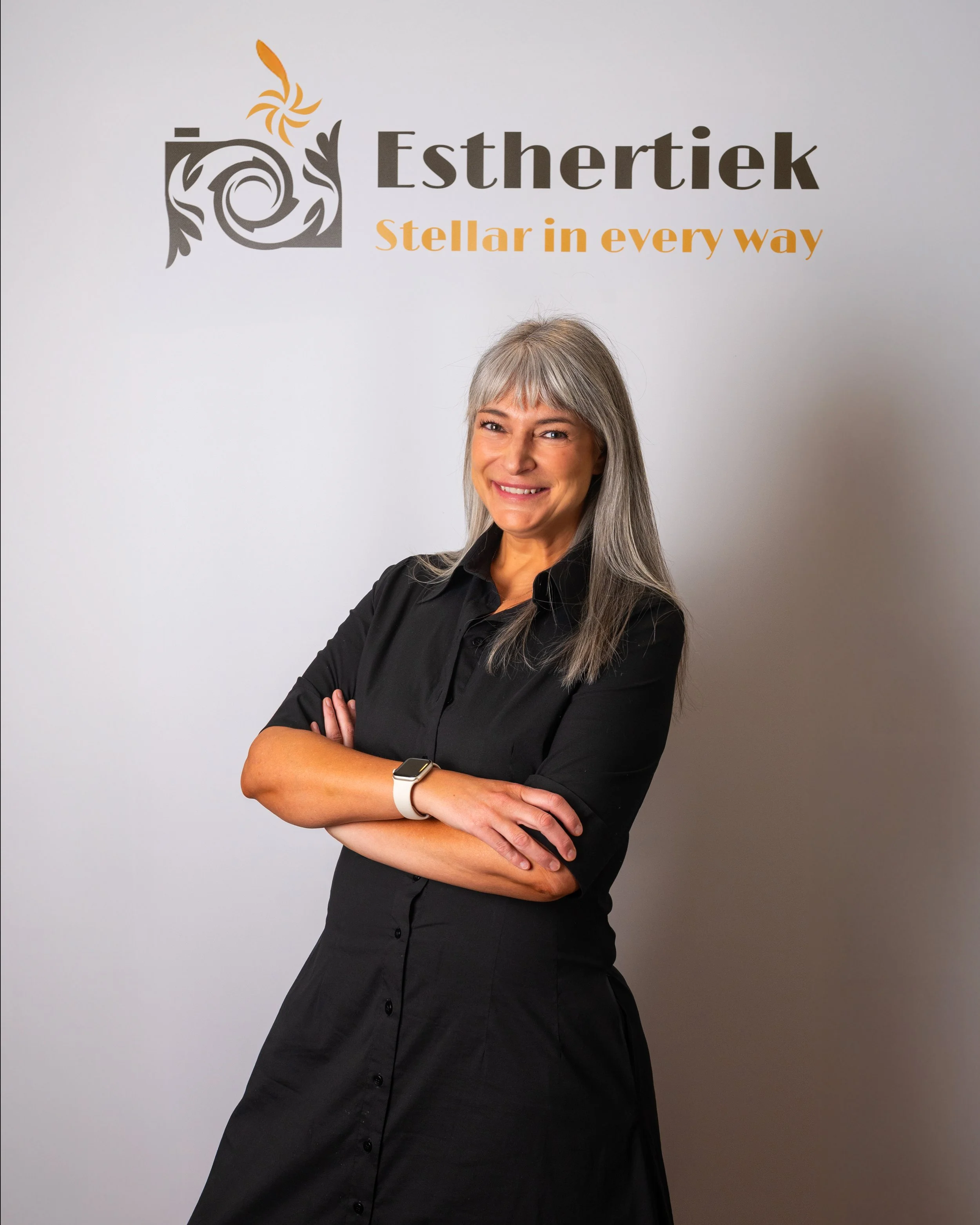 A woman with gray hair, wearing a black dress and a smartwatch, standing with arms crossed in front of a white wall with the company logo 'Esthertiek' and the tagline 'Stellar in every way'.
