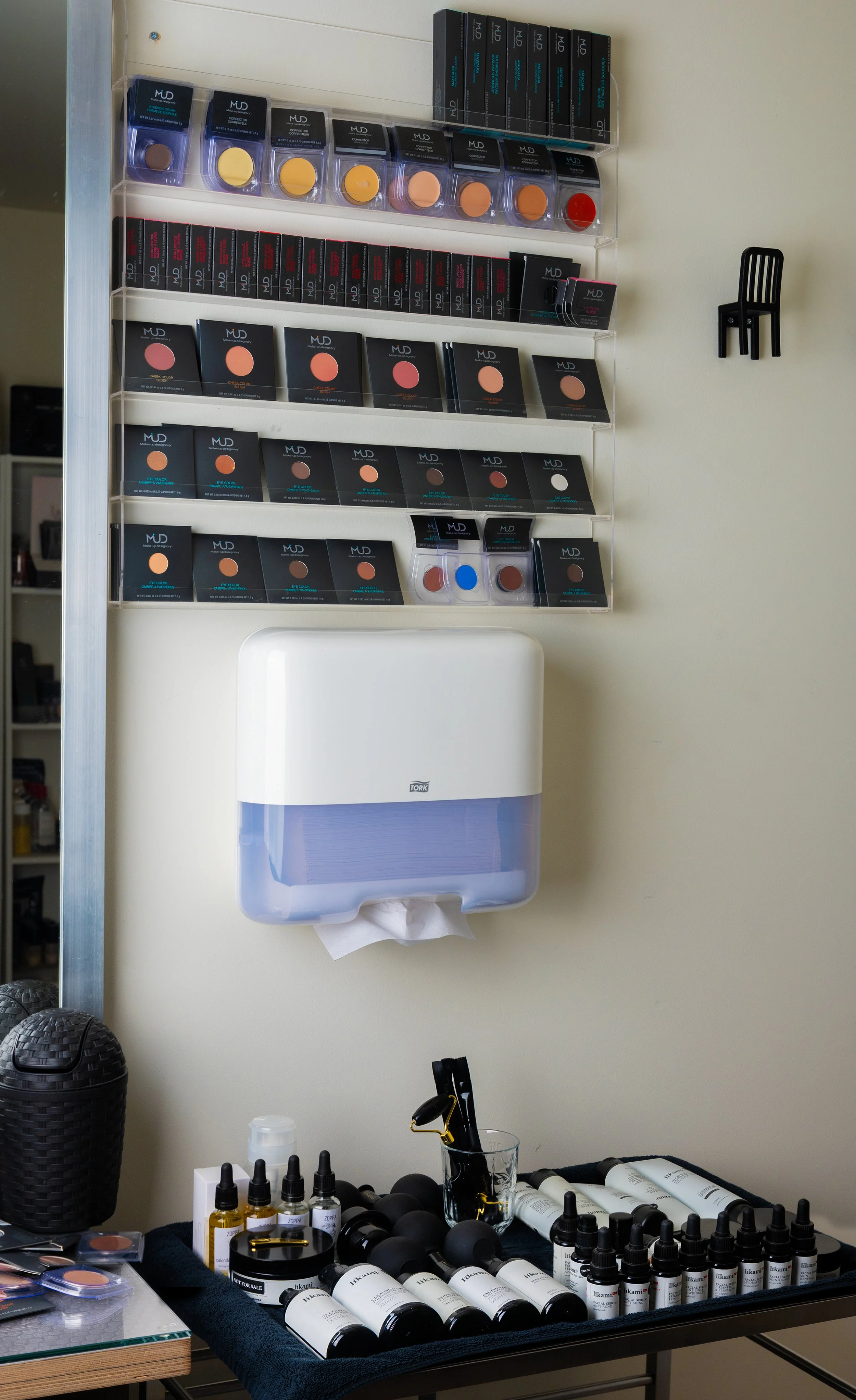 Display of makeup products, including various shades and types of cosmetics, with a tissue dispenser on the wall and makeup tools on a table.