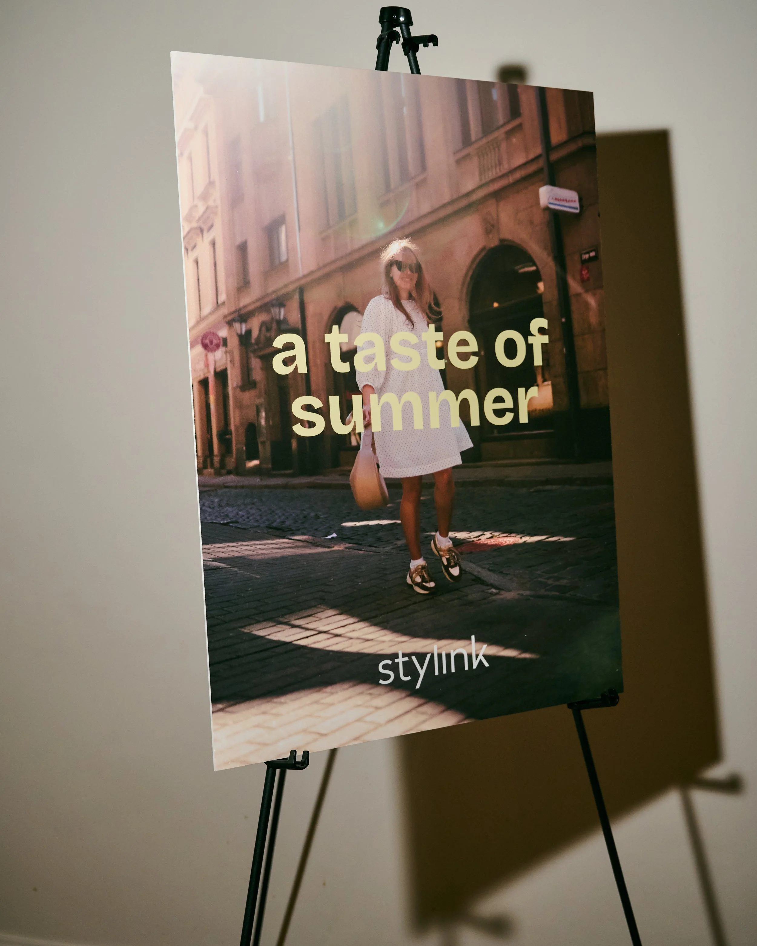 Poster featuring a woman in sunglasses standing on a city street with old buildings in the background, with the words "a taste of summer" and the logo "stylink" at the bottom.