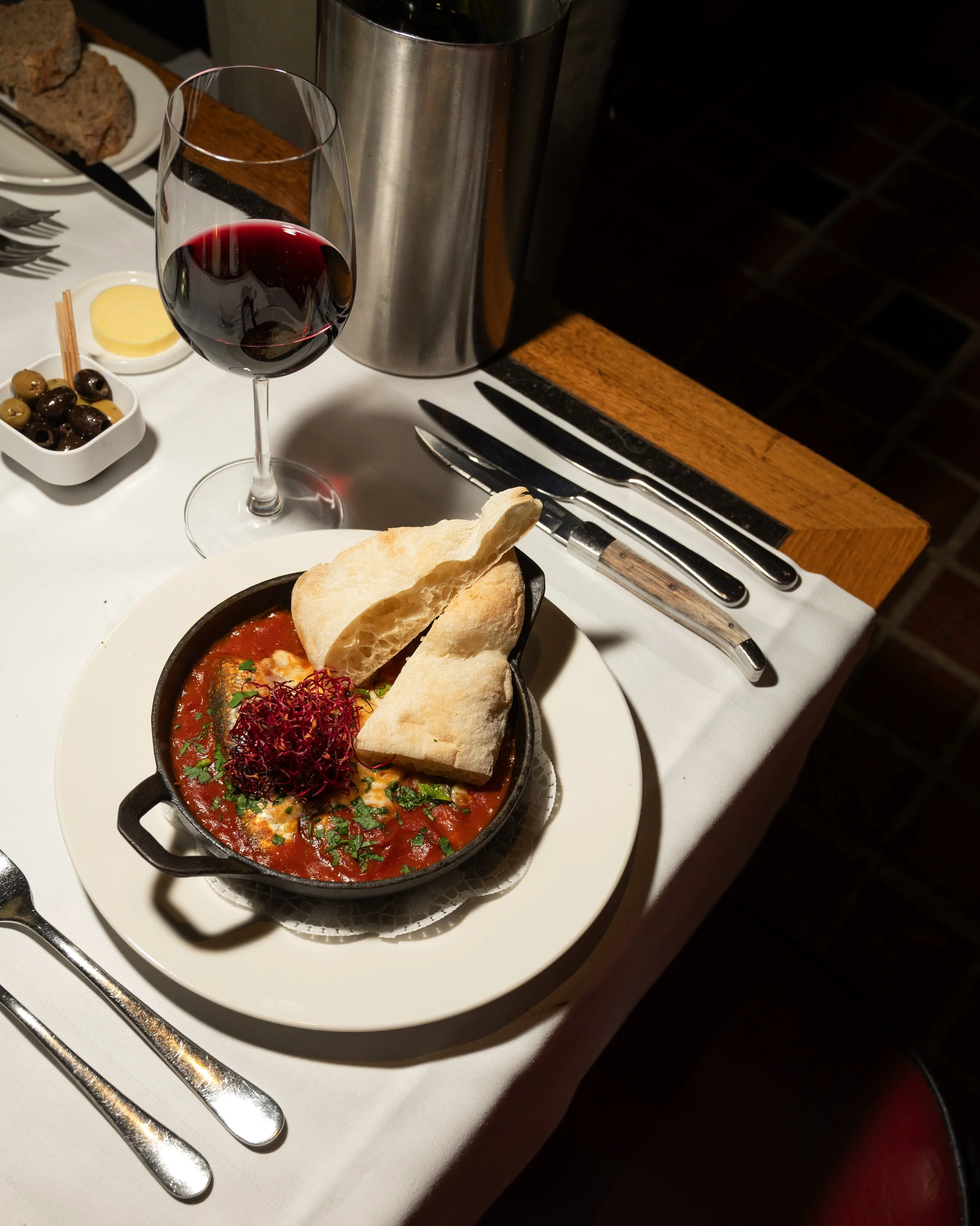 A table setting featuring a bowl of stew with bread, a glass of red wine, and side dishes, with silverware and a white tablecloth.