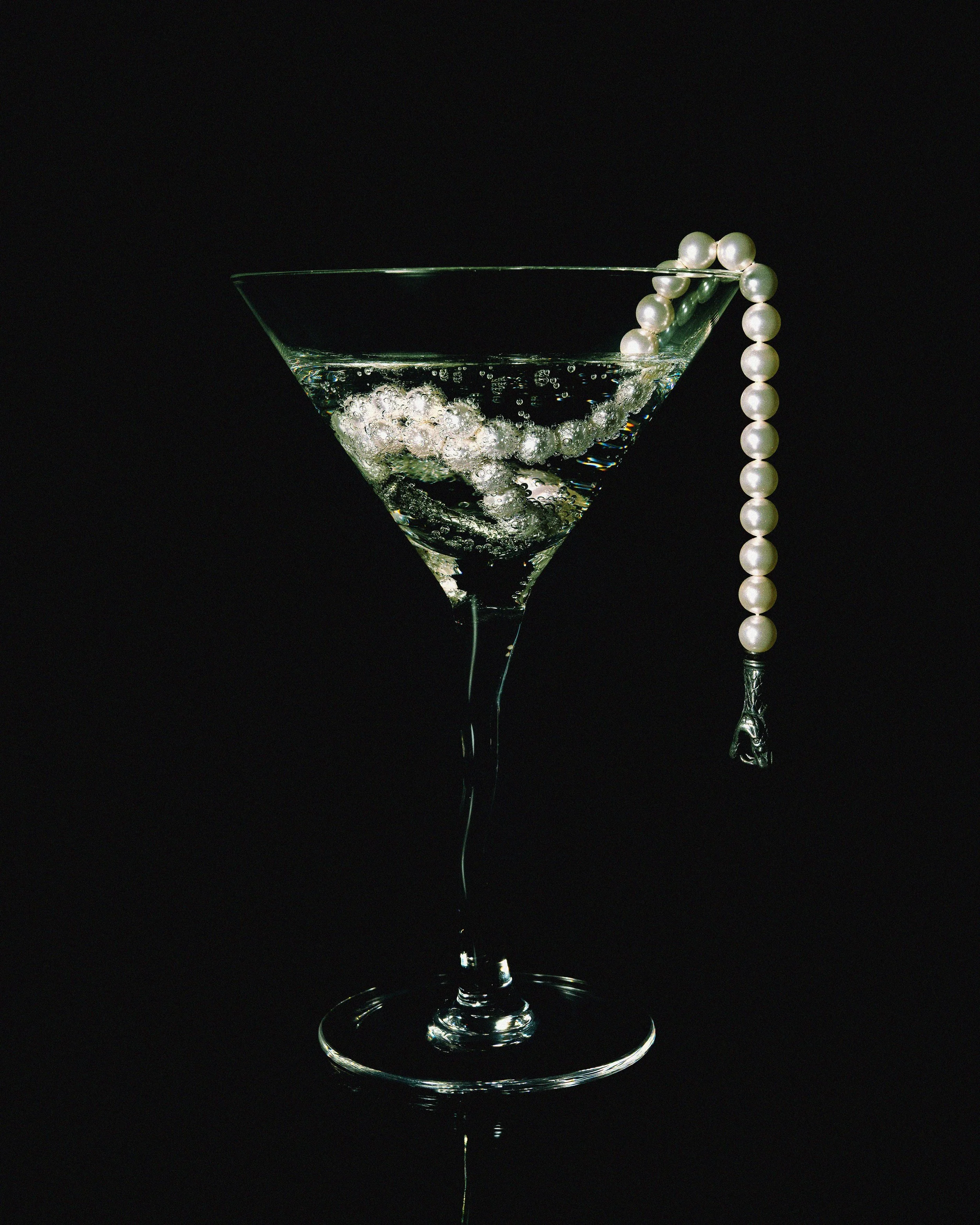 A martini glass filled with a clear liquid, bubbles rising within, garnished with a string of white pearls draped over the rim, against a black background.