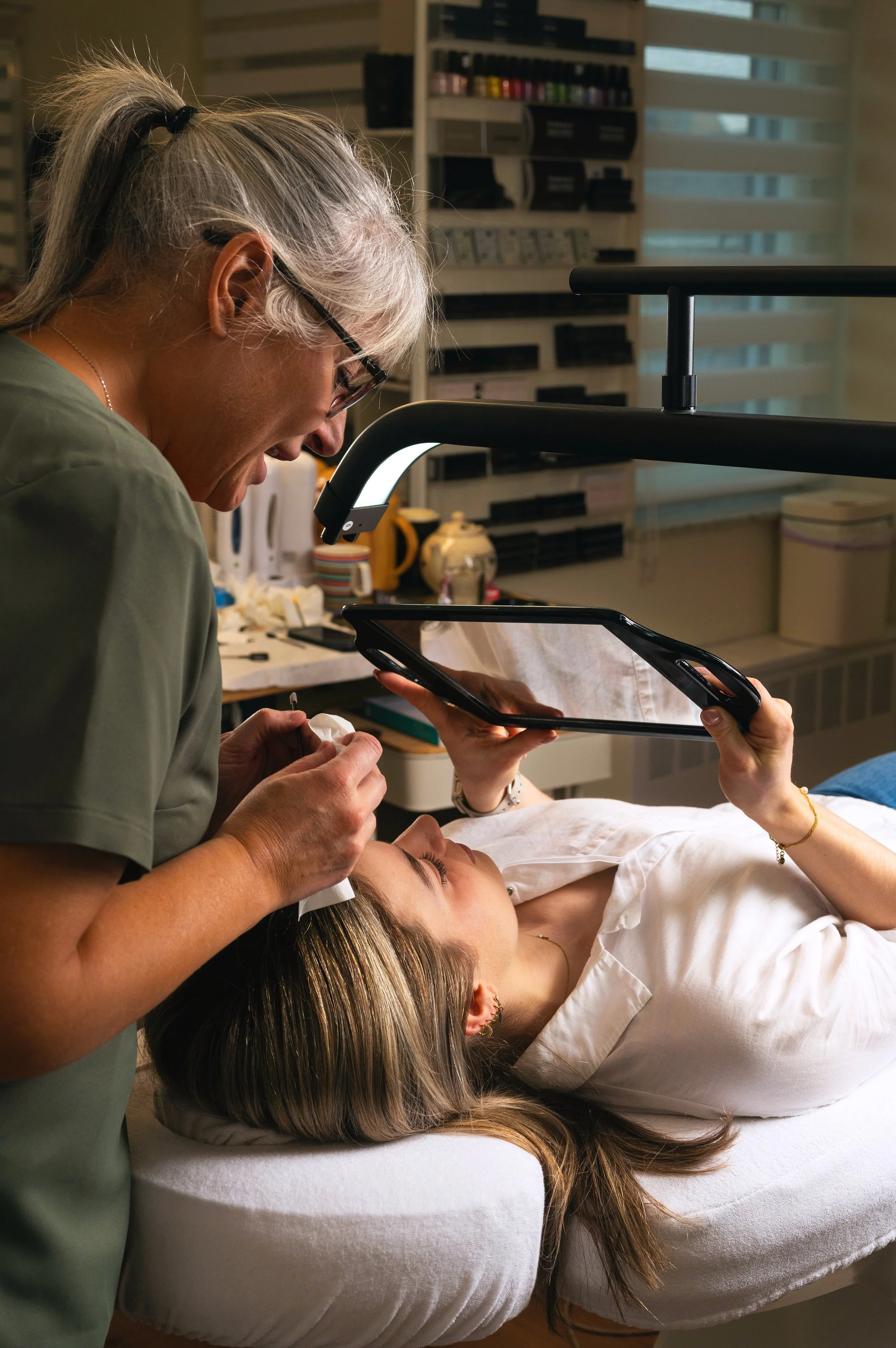 A healthcare professional performs a cosmetic procedure on a woman lying on a spa bed, with a magnifying mirror and medical tools.
