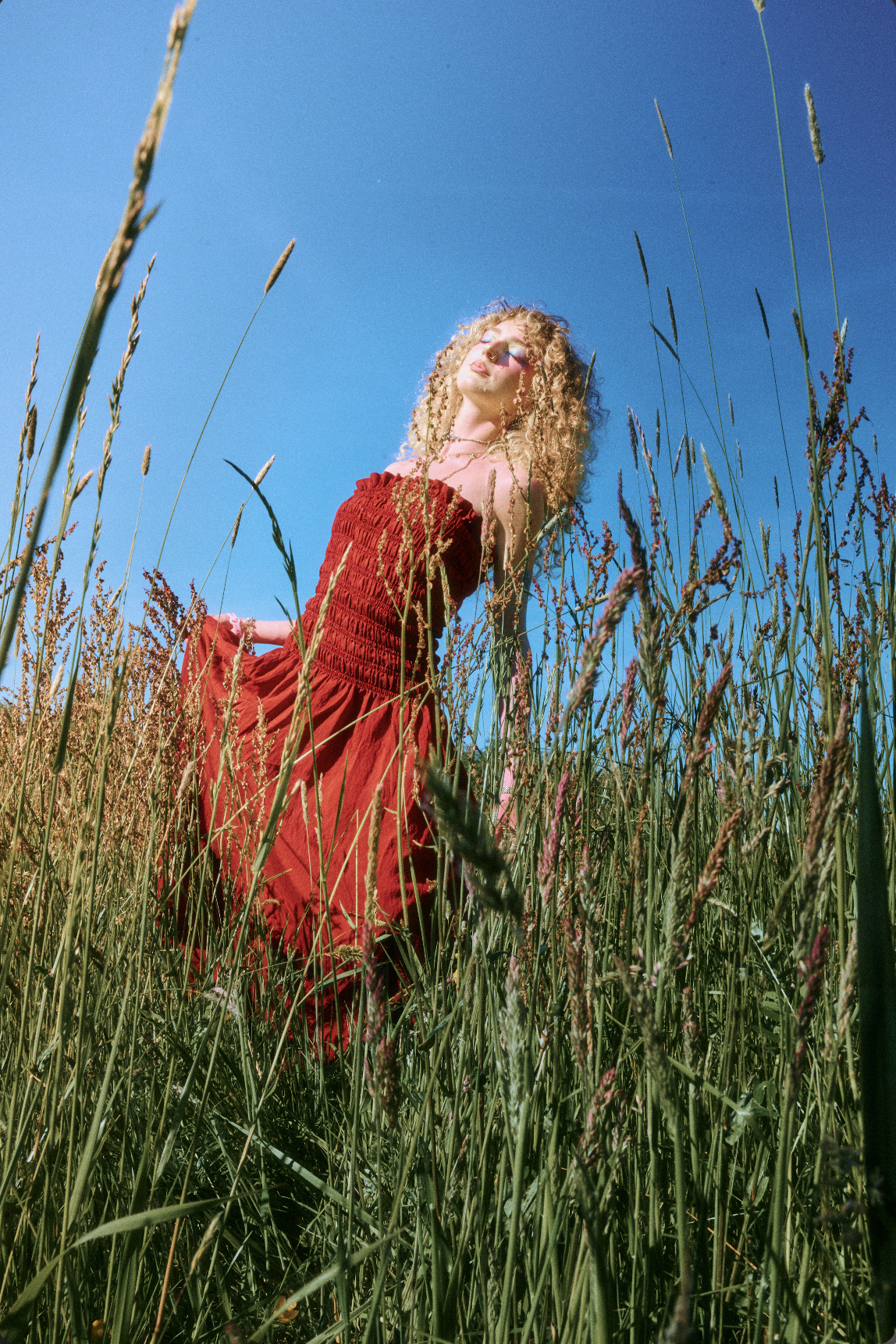 A woman with curly blonde hair standing in a tall grassy field wearing a red strapless dress with her eyes closed and face tilted upward