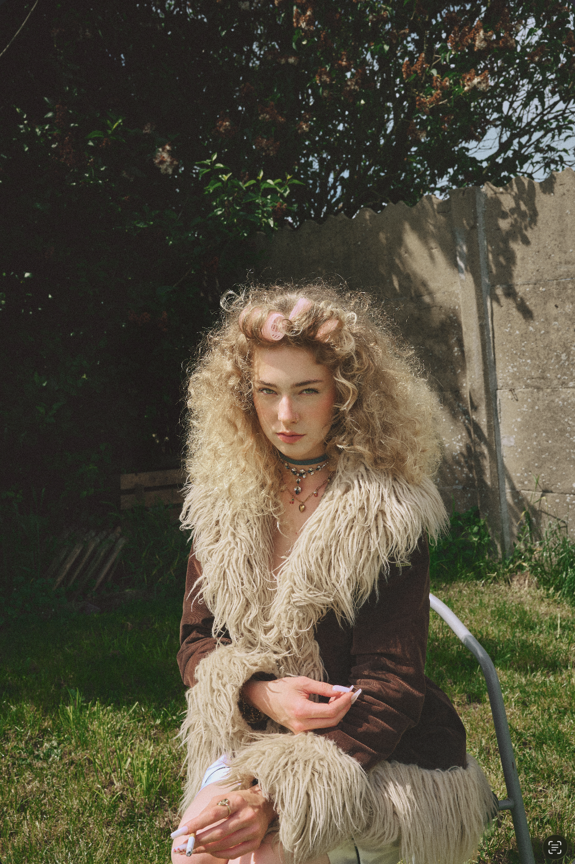 A young woman with curly blonde hair and a serious expression, wearing a fluffy beige vest over a brown jacket, seated outdoors on a metal chair with green grass, plants, a concrete wall, and a bush in the background.