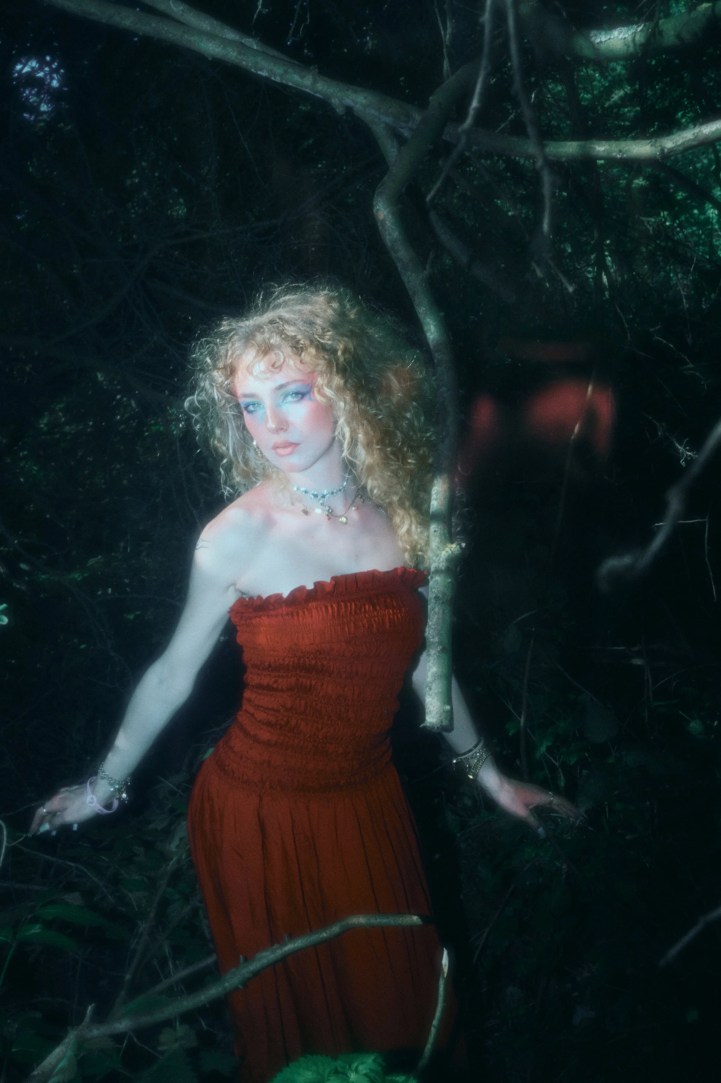 A young woman with curly blonde hair and blue eye makeup, wearing a strapless red dress, is standing in a forest surrounded by dark branches and leaves.