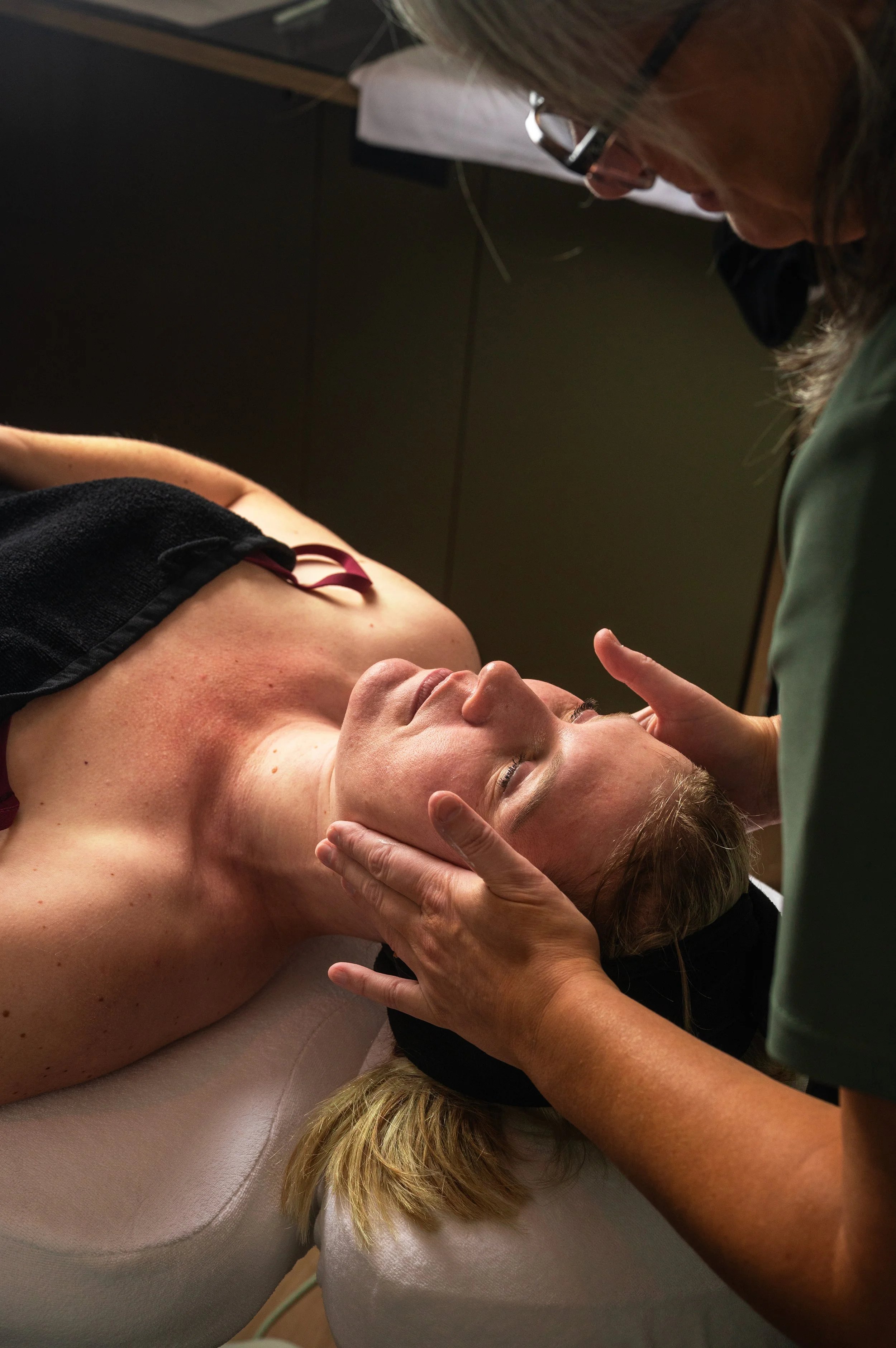 A woman receiving a chiropractic or massage therapy treatment, lying on her back on a treatment table while a healthcare professional gently holds her head and neck.