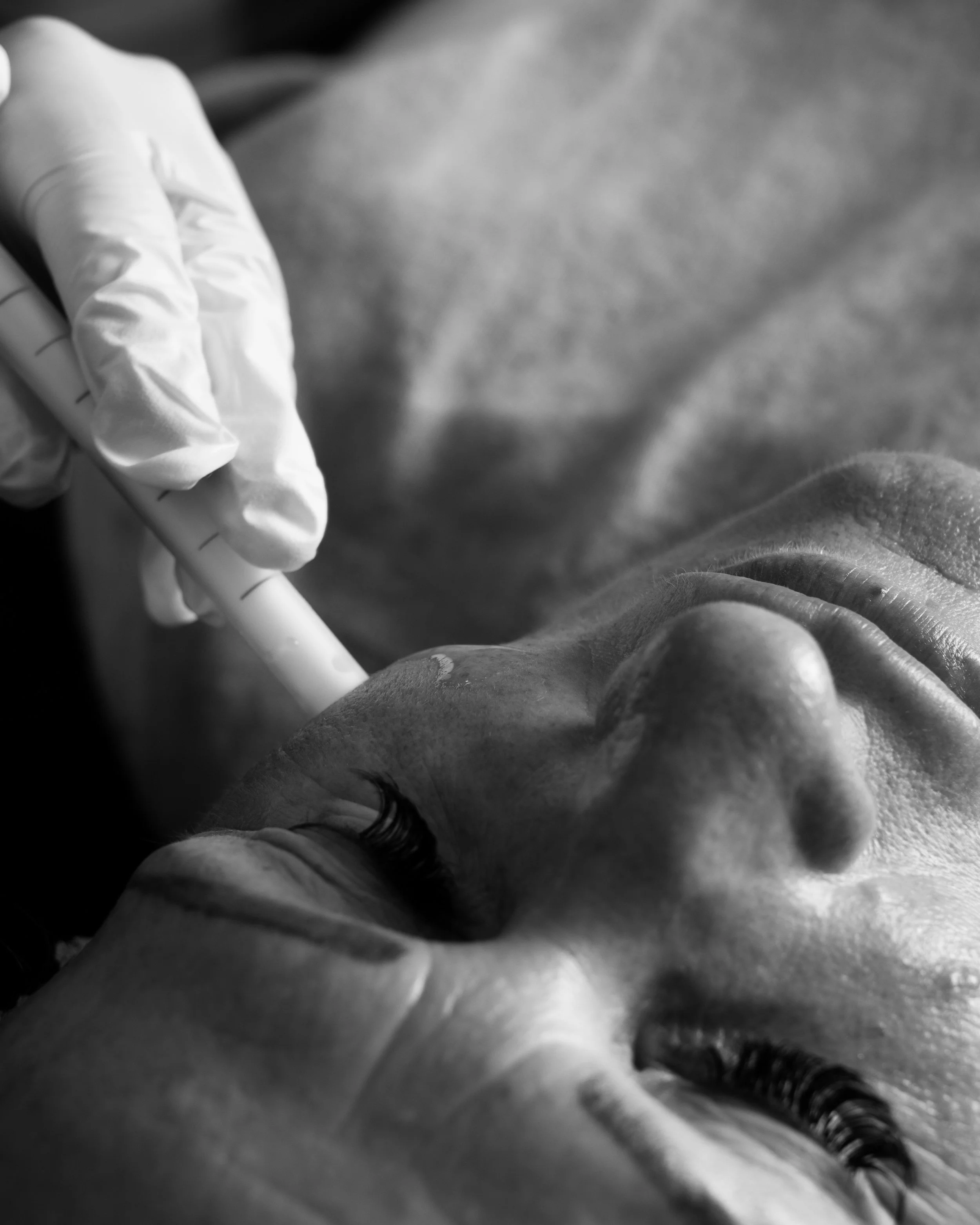Close-up black and white photo of a person in medical gloves holding a syringe, possibly preparing for an injection, with blurred background.