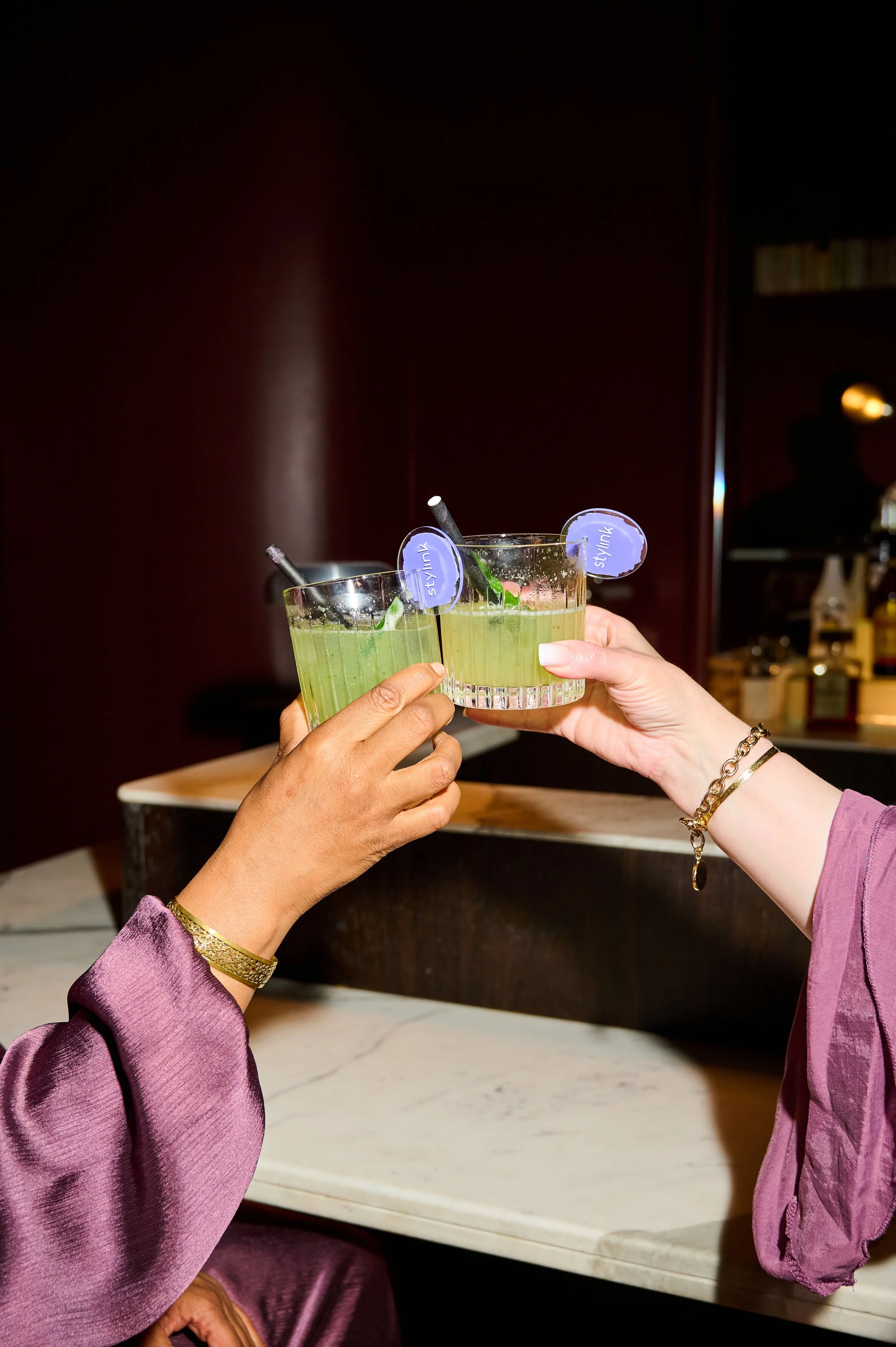 Two people are clinking glasses filled with a green beverage, garnished with lime and a purple tag labeled 'styling'. The scene is indoors with a dark background and a person in silhouette in the distance.