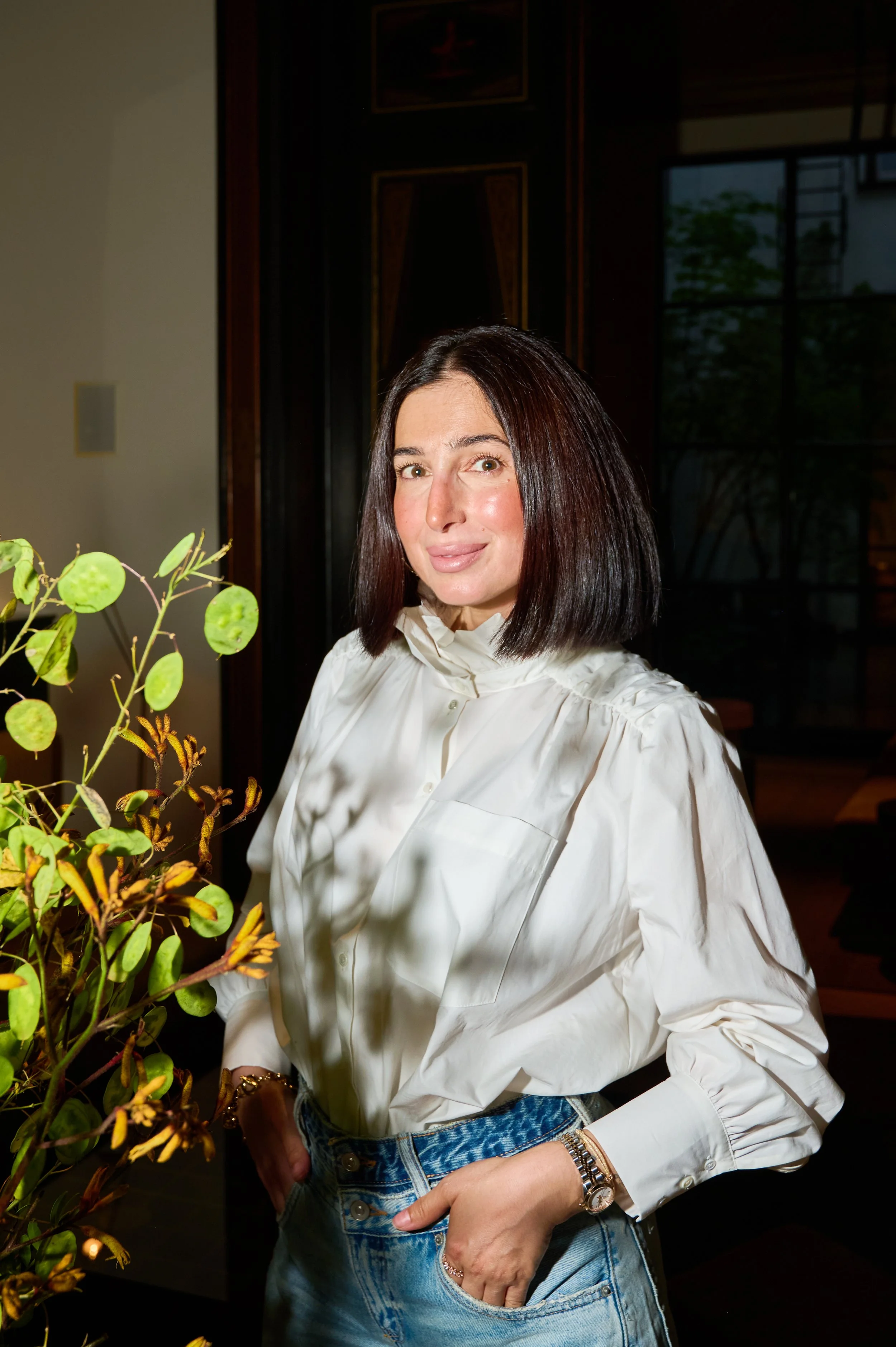 A woman with shoulder-length dark hair wearing a white button-up shirt and denim jeans, standing indoors near a plant with green and yellow leaves, with a large window and dark wood paneling in the background.