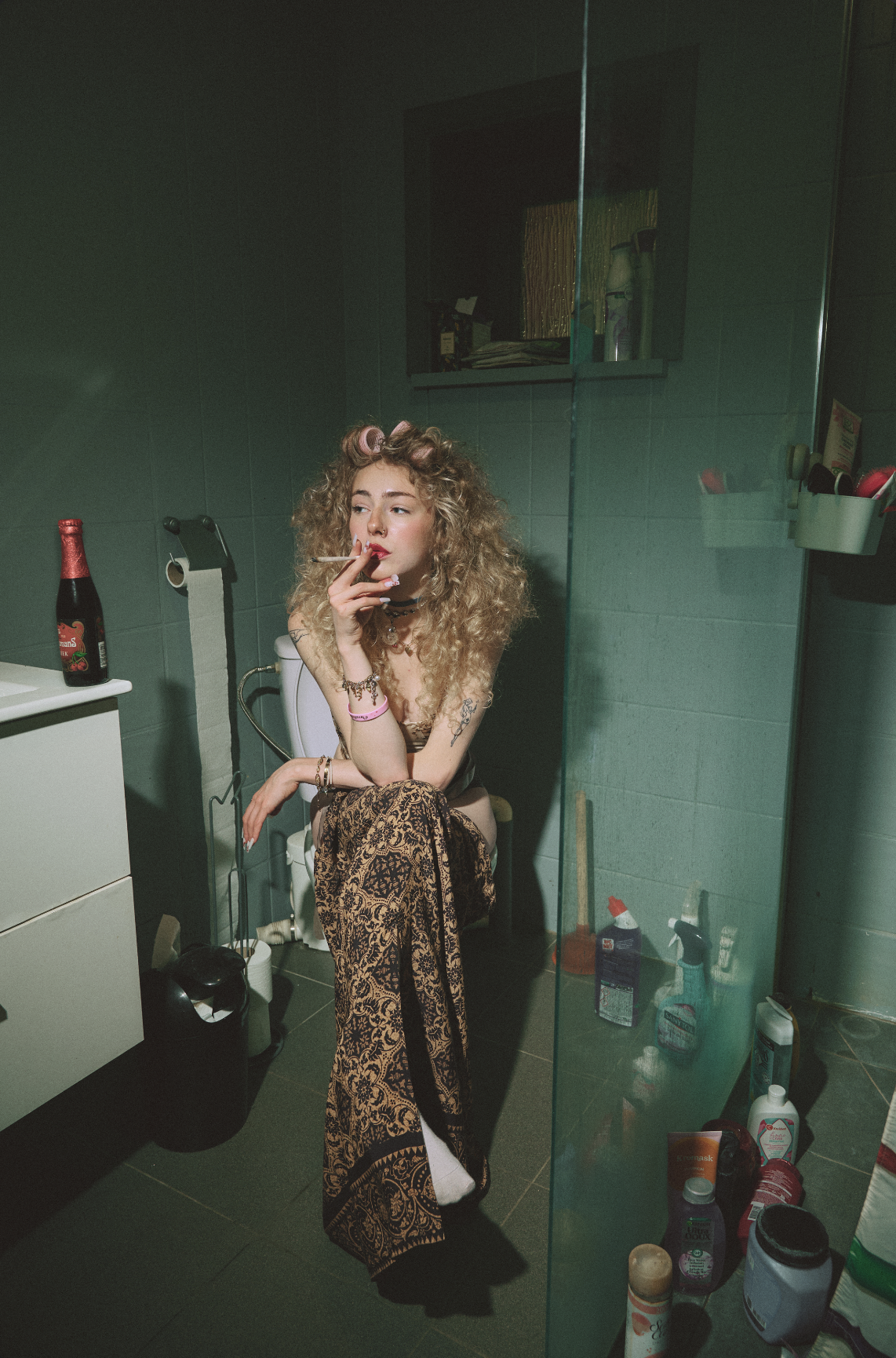 A young woman with curly blonde hair, tattoos, and wearing patterned pants, is sitting on a toilet in a bathroom. She has curlers in her hair, is smoking a cigarette, and has various jewelry and wristbands. The bathroom has green tile walls, a glass 