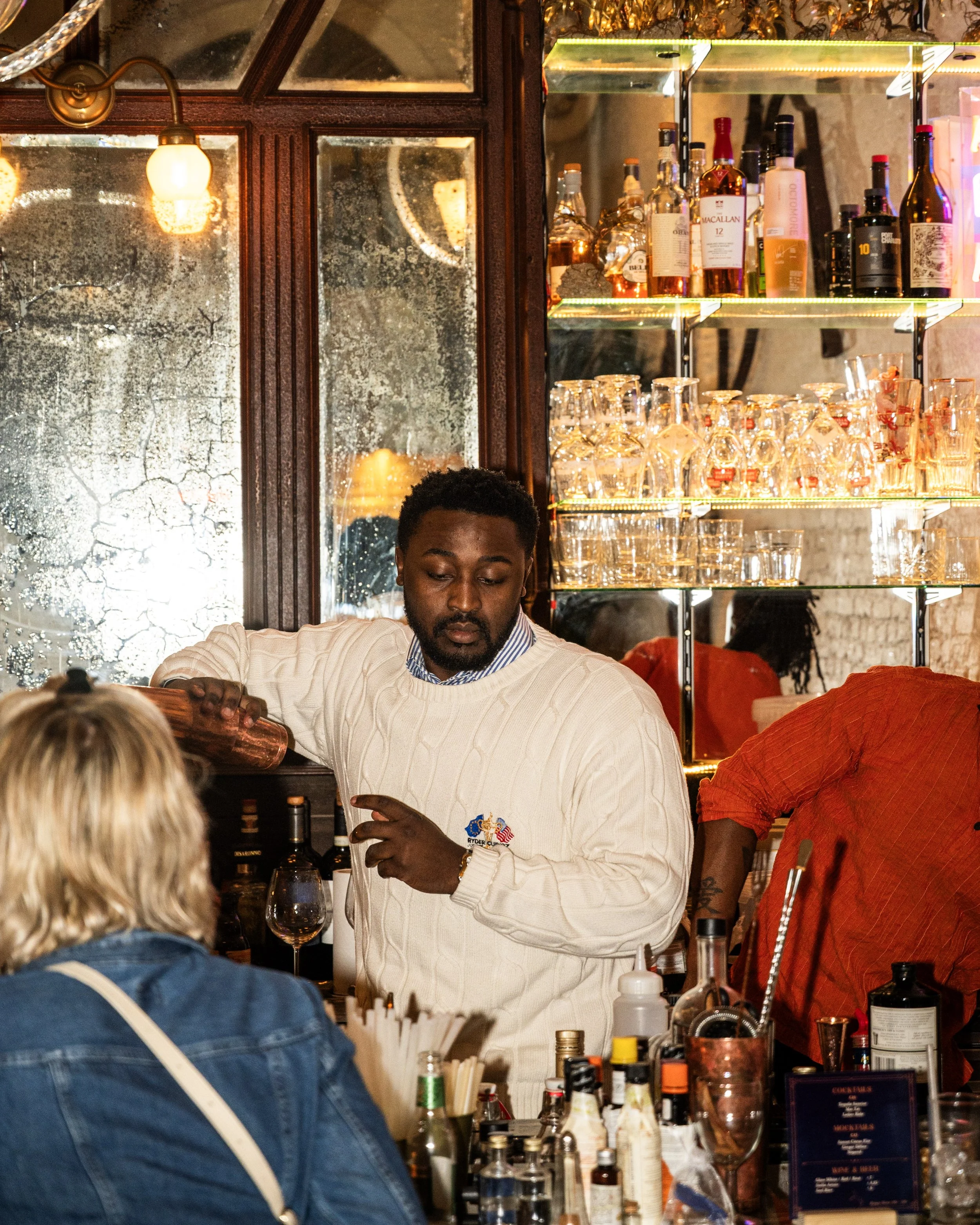 A bartender with dark hair and a beard, wearing a white sweater, serves a woman with blonde hair in a denim jacket at a bar. The bar has bottles, mixers, and glasses, with a glass shelf behind the bartender displaying liquor bottles and glassware. Th