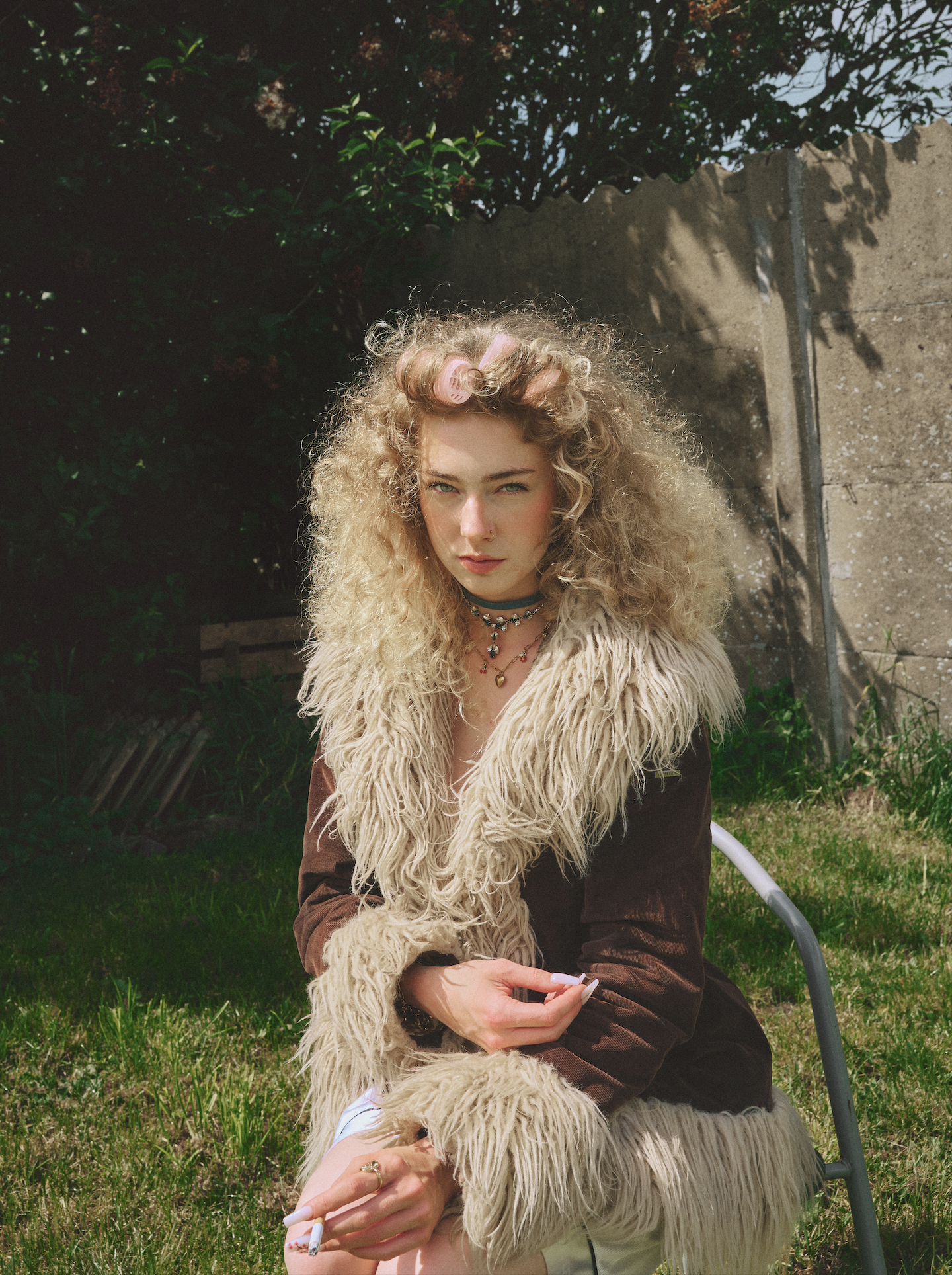 Young woman with curly blonde hair in curlers, wearing a brown jacket with a furry collar, sitting outside on the grass holding a cigarette, with green plants and a concrete wall in the background.