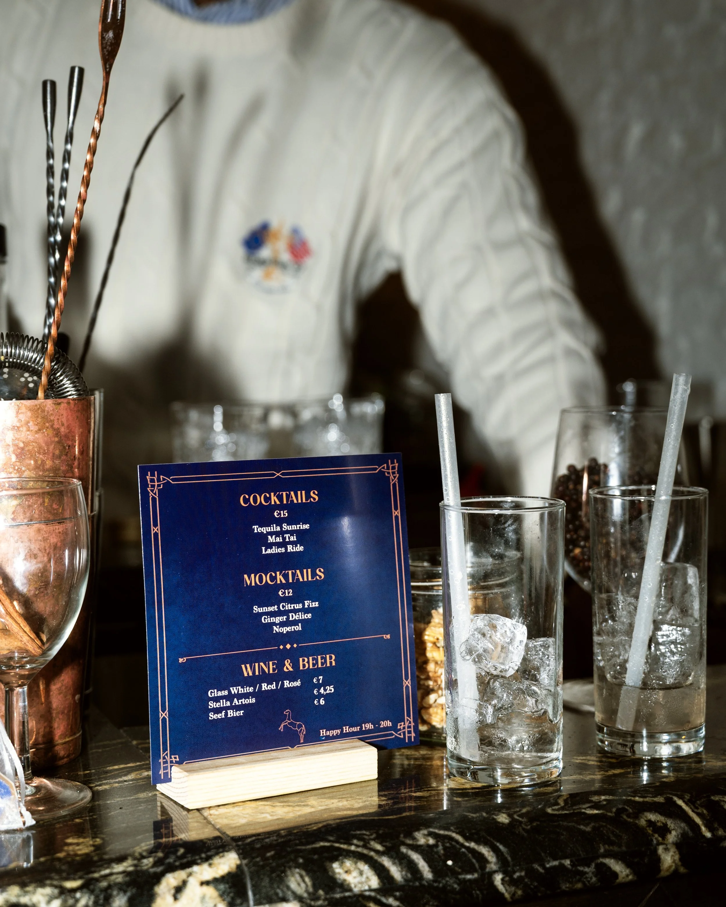 Bar counter with a blue menu, various glasses with straws and ice, and bartender in a white shirt in the background.