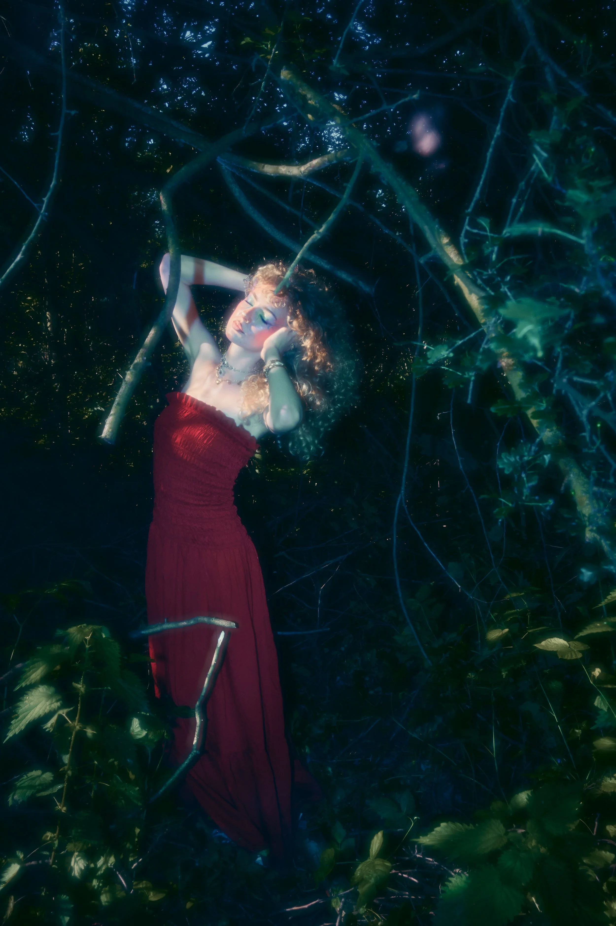 A woman with curly hair, wearing a red strapless dress, is surrounded by dense green foliage and branches, with her eyes closed and her hands gently touching her head in a serene pose.