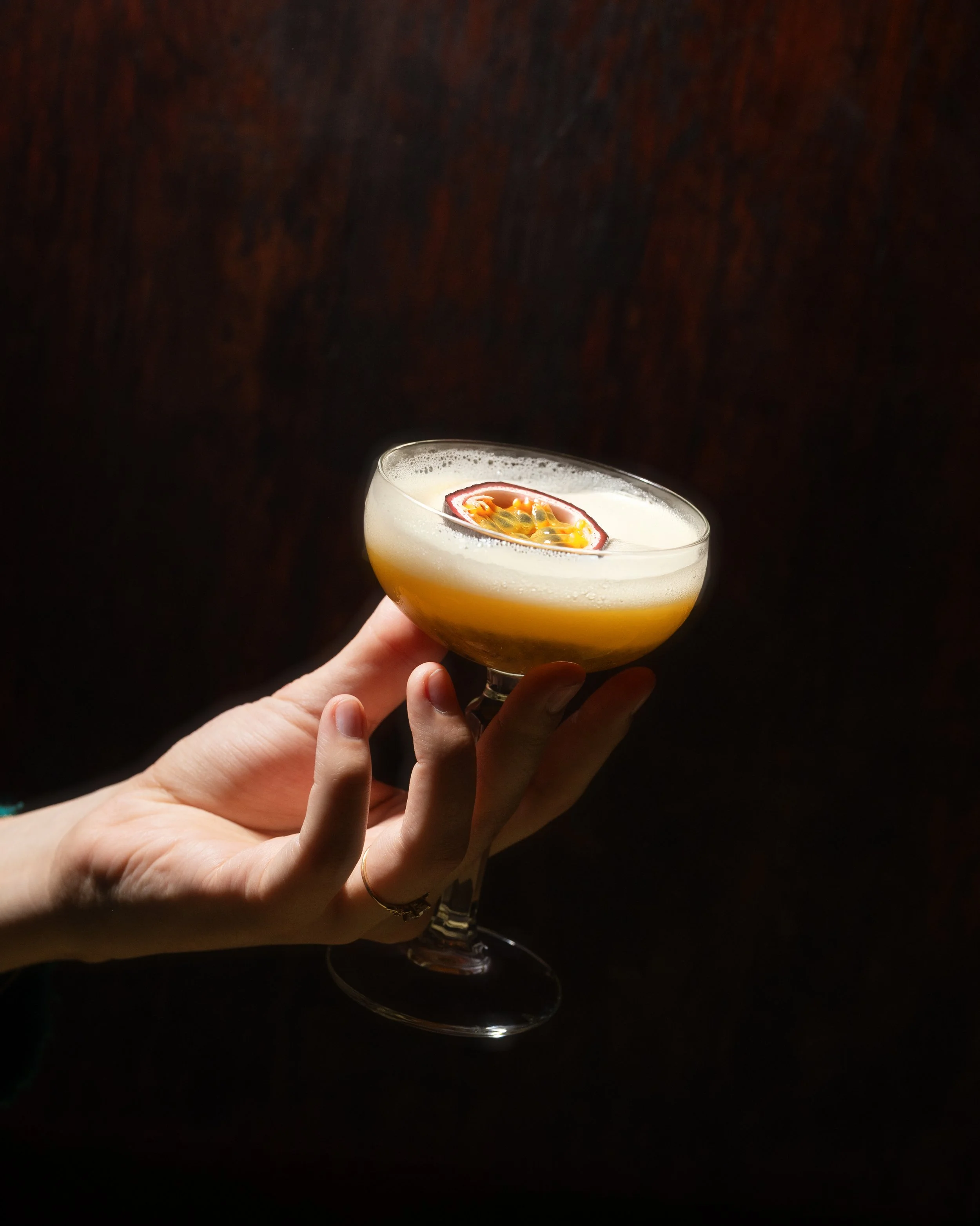 A hand holding a cocktail glass with a yellowish drink and a passion fruit garnish.