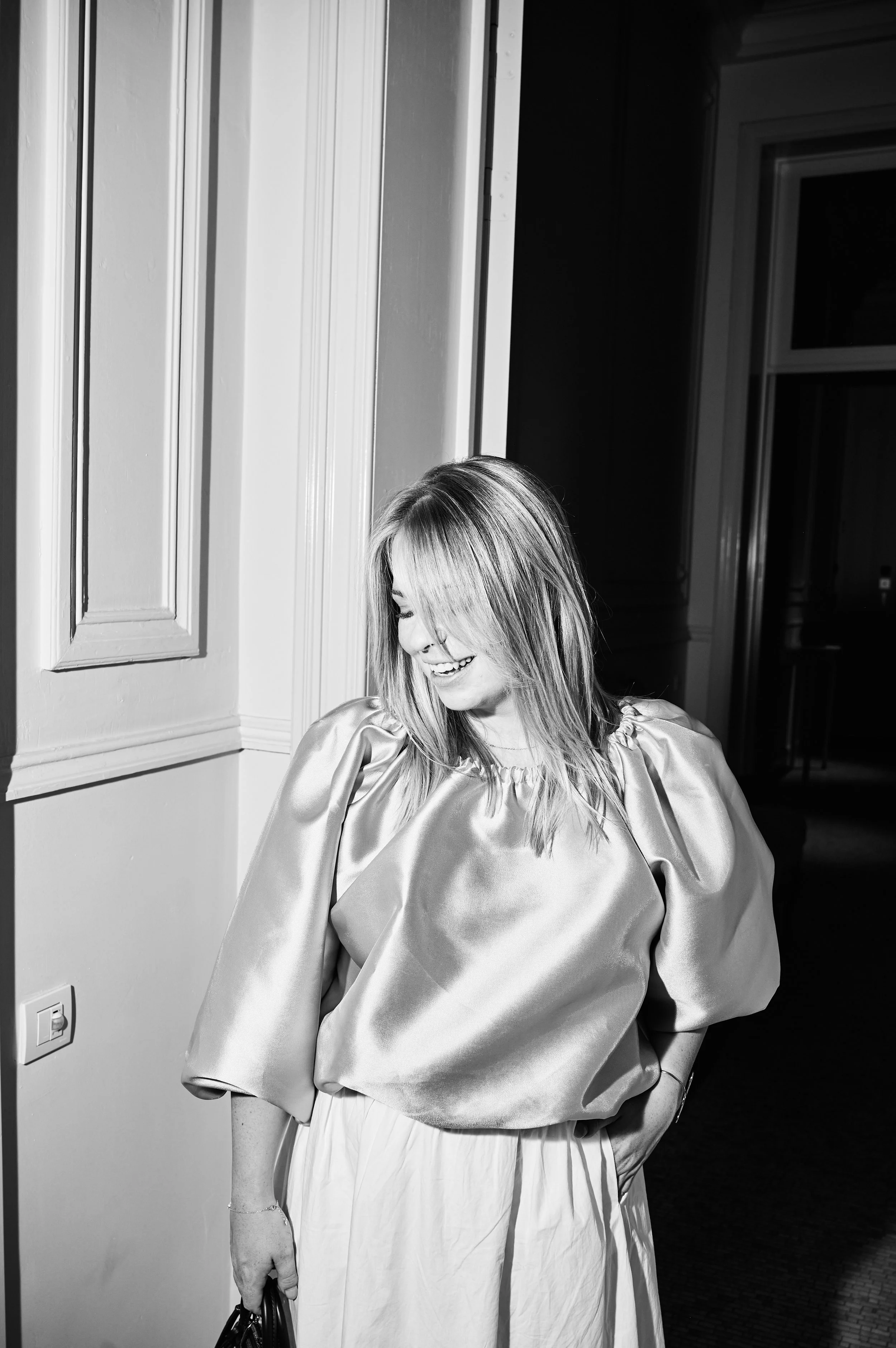 A woman with shoulder-length hair, smiling and looking down, wearing a shiny satin or silk dress with puffed sleeves and a pleated skirt, standing indoors near a wall with framed panels and a window in the background.