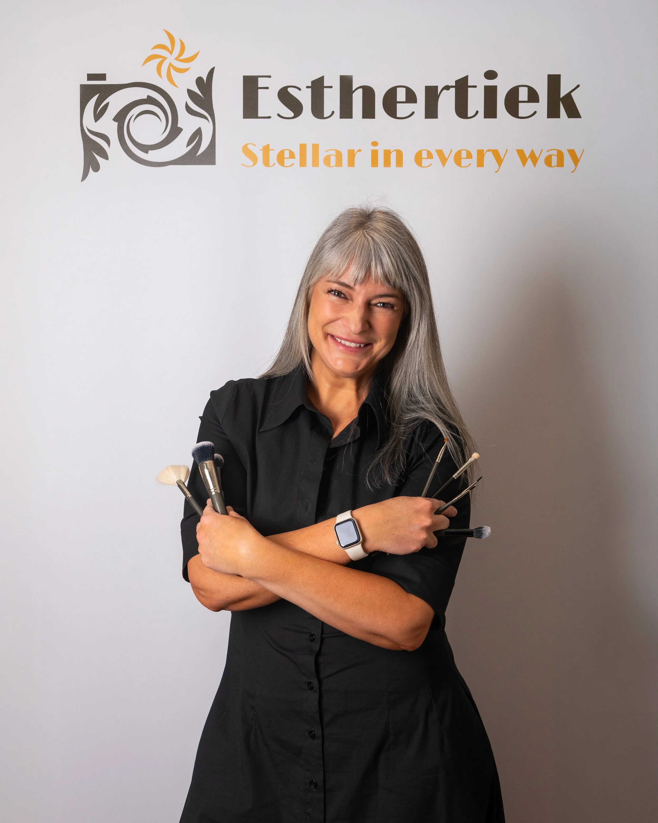 A woman with gray hair smiling, holding makeup brushes, standing in front of a wall with the logo 'Esthertiek' and the slogan 'Stellar in every way.'