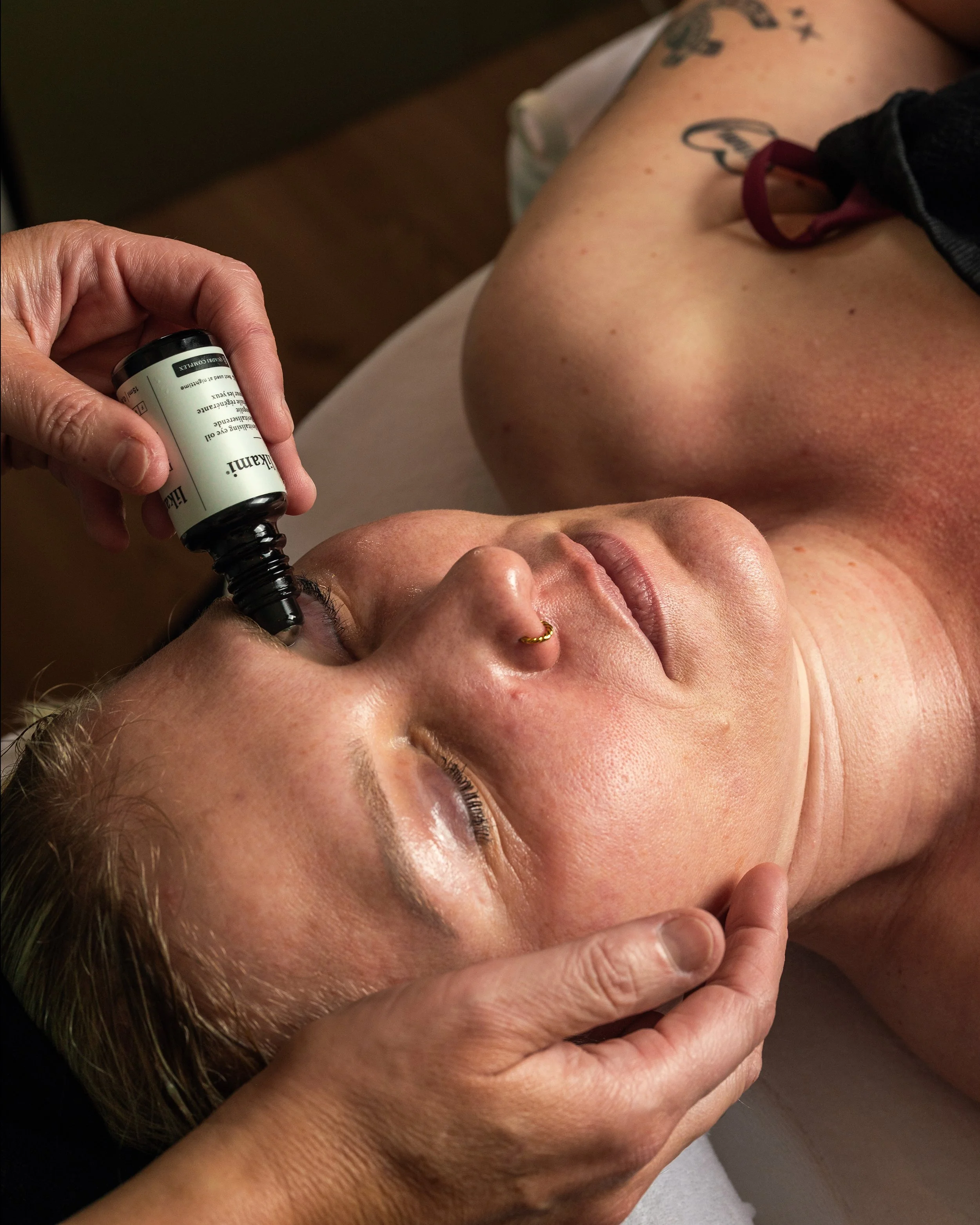 A woman with a nose piercing receives a facial treatment using a black dropper from a bottle, lying on a spa bed with eyes closed.