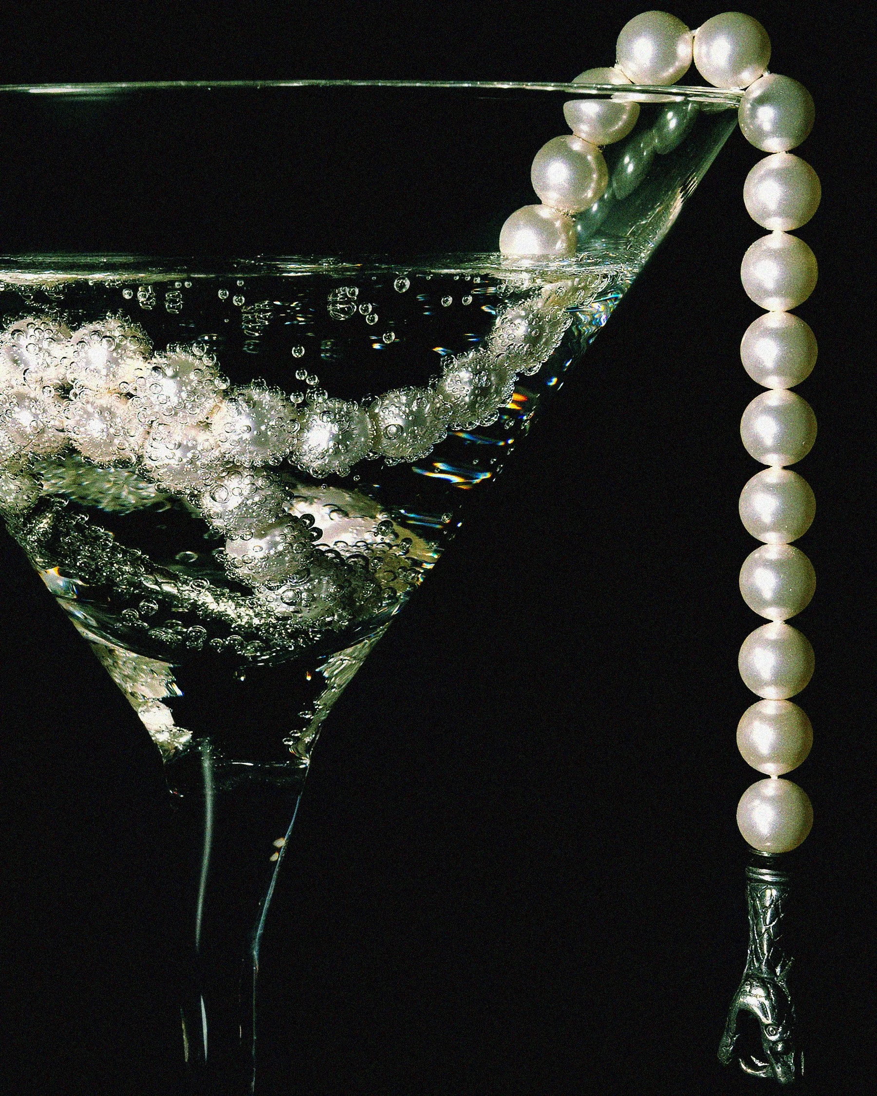 A martini glass with pearls draped over the rim, set against a black background, with a string of pearls beside it.