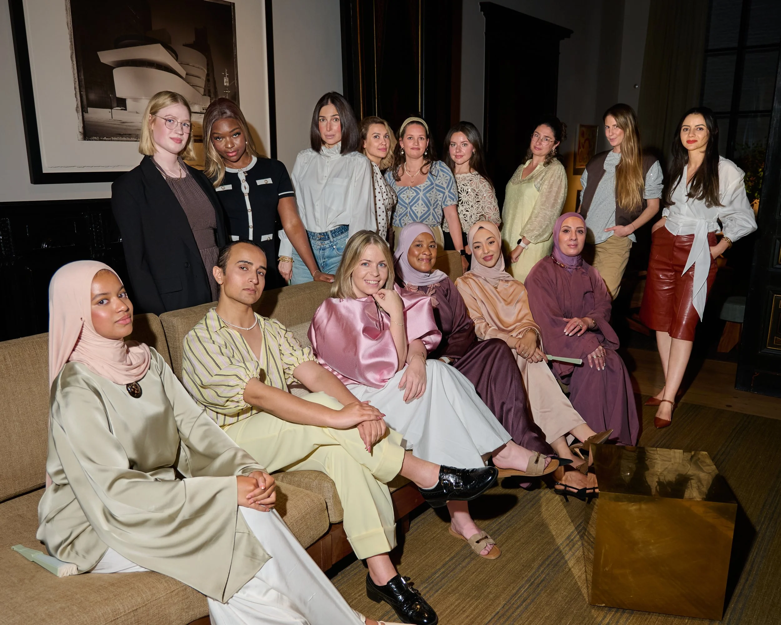 Group of women sitting and standing in a room with dark walls, framed pictures, and a window, some wearing traditional clothing and others in contemporary outfits.