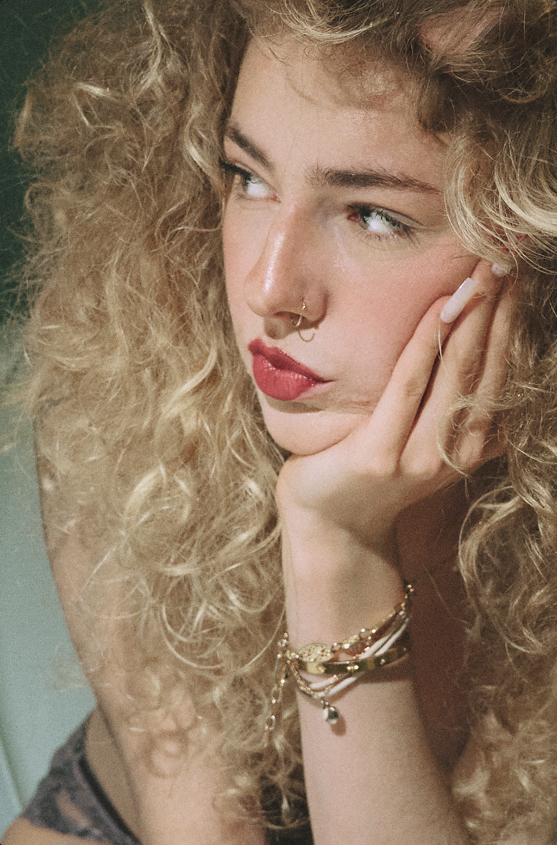 A young woman with curly blonde hair, red lipstick, and a nose ring, resting her face on her hand and looking to the side.