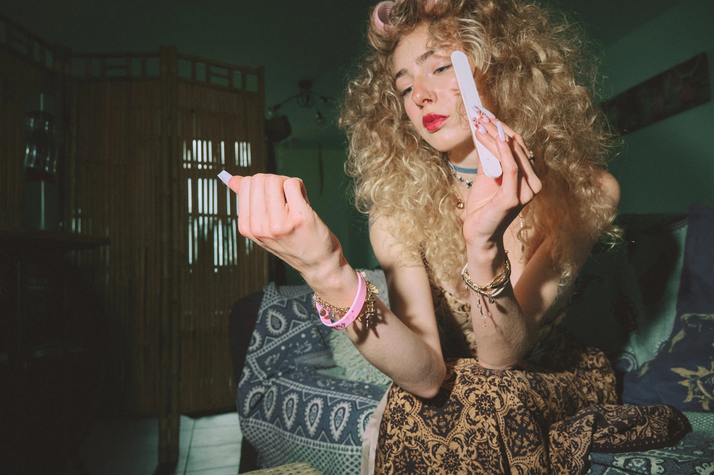 A woman with curly blonde hair, red lipstick, and various jewelry holding a nail file while sitting in a room with wooden decor.