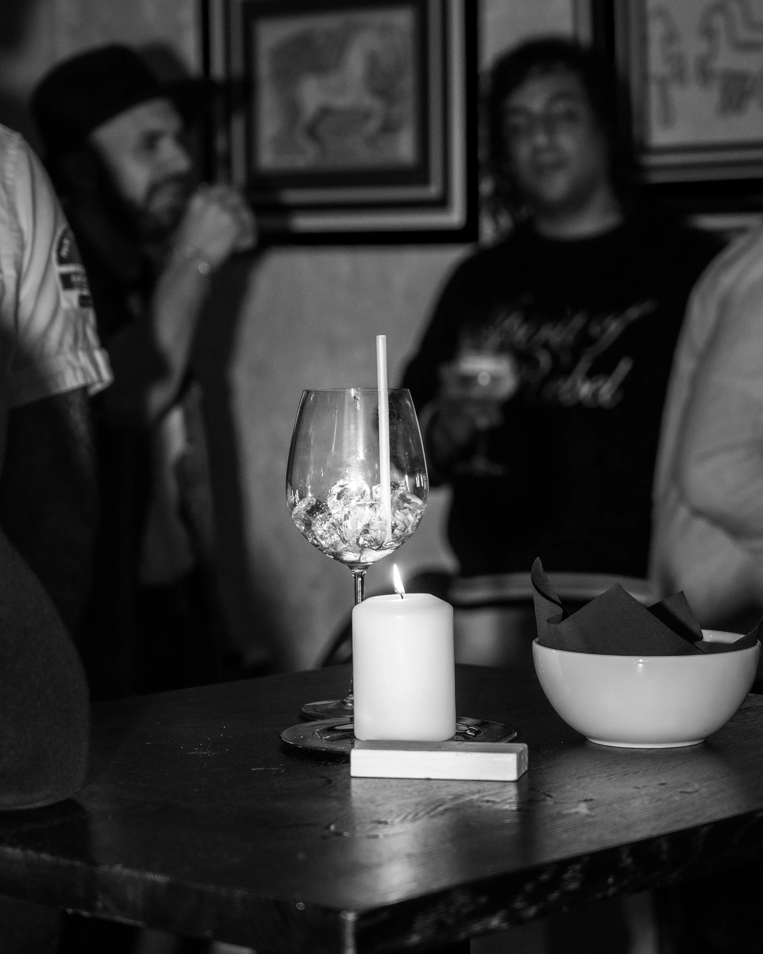 A table with a candle, a glass with ice and a straw, and a bowl with napkins in a dimly lit setting, with people in the background at a social gathering.