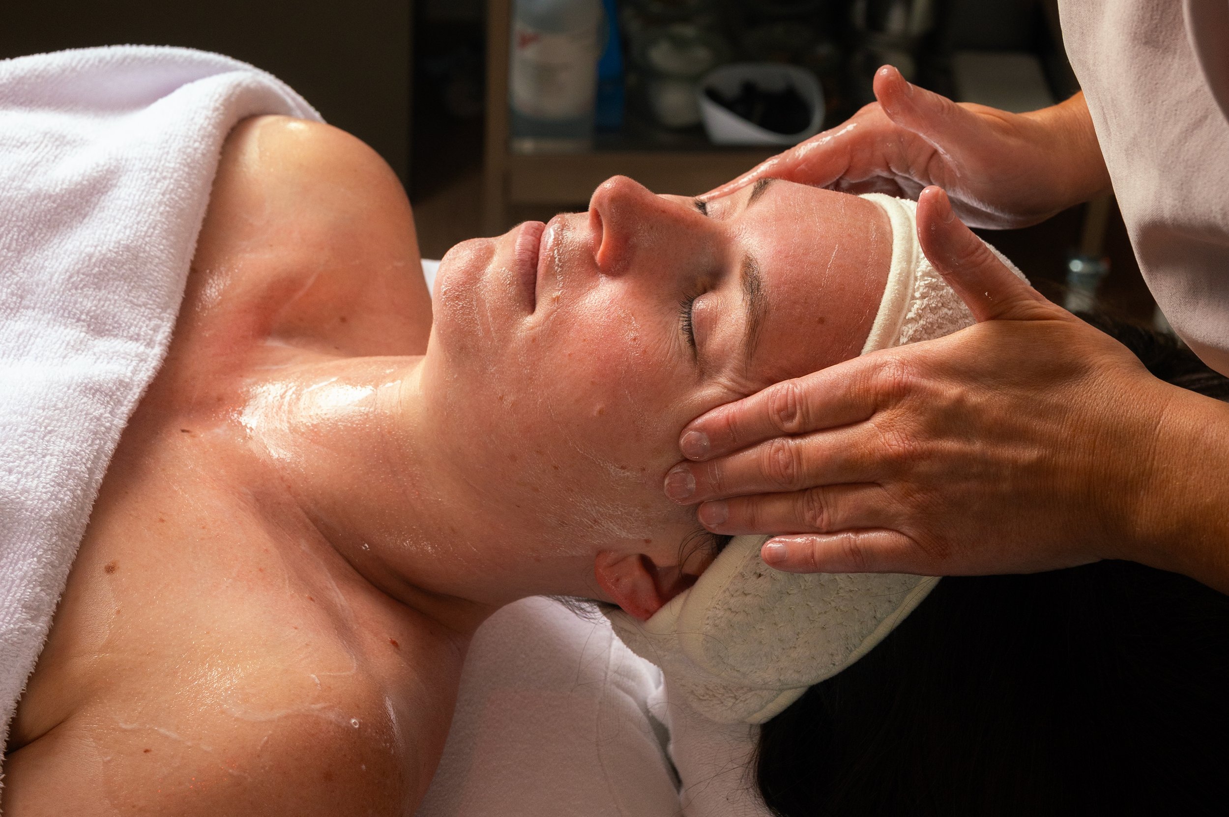 A woman receiving a facial massage or treatment at a spa, lying on her back with eyes closed, a towel covering her chest, and a headband around her forehead.