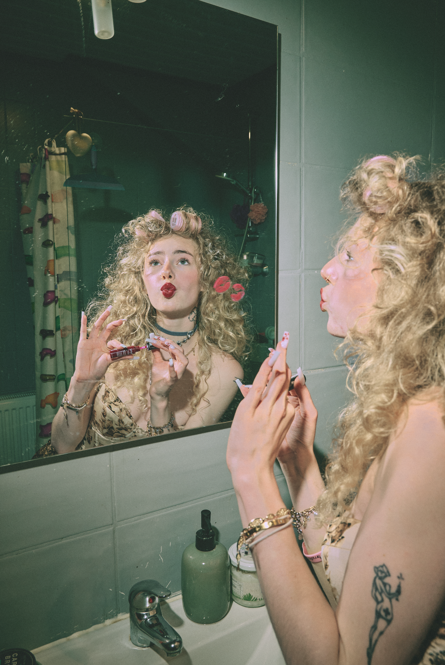 A woman with curly blonde hair and tattoos is looking at herself in a bathroom mirror, holding a lipstick and applying makeup. The mirror reflects her face and part of her upper body, with a heart-shaped decoration and curtain visible in the backgrou