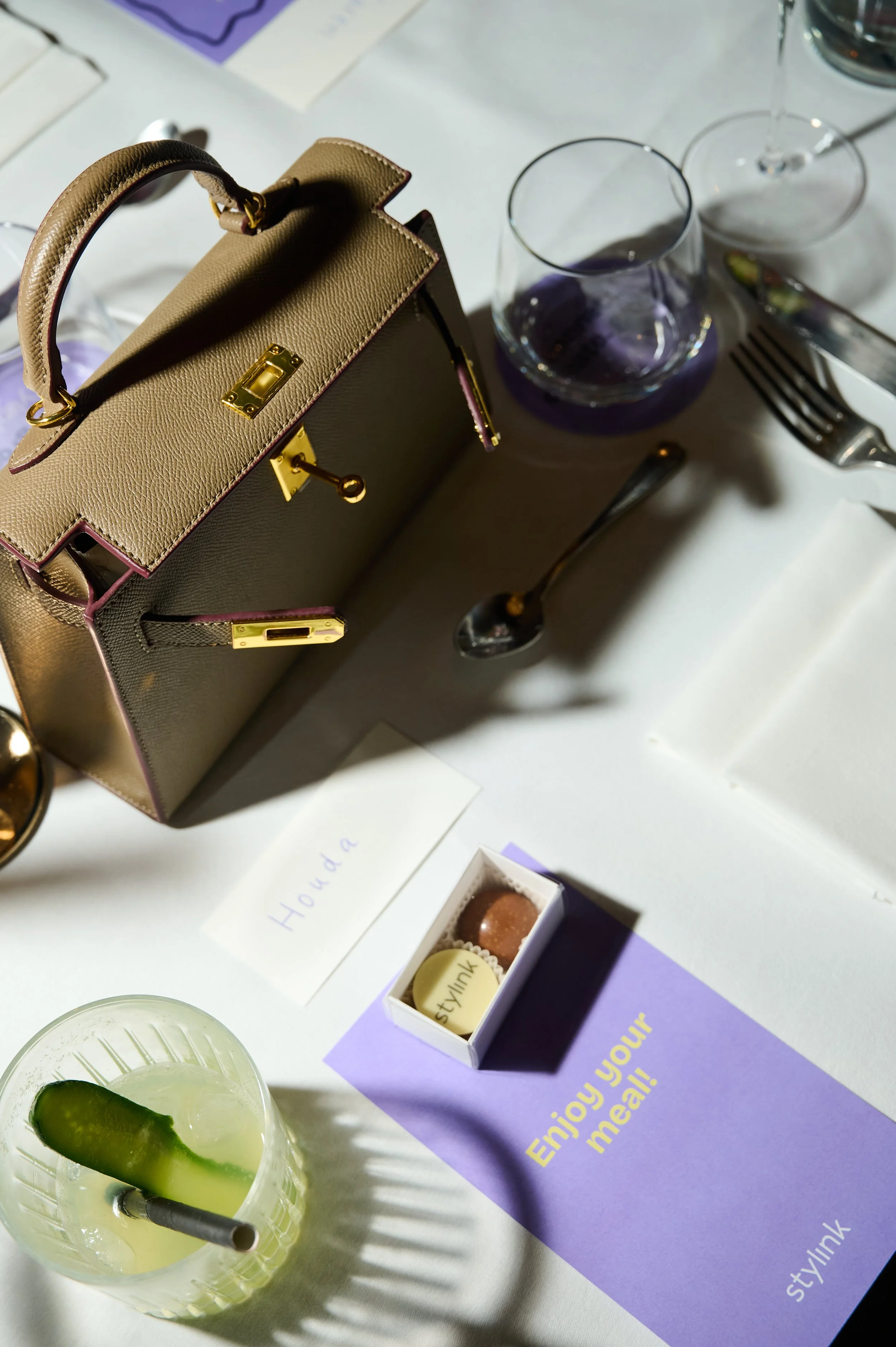 A beige handbag with gold hardware on a table with various items including glasses of water, a fork, and a purple paper with yellow text, along with a small box of chocolates and a drink garnished with a cucumber slice and a straw.
