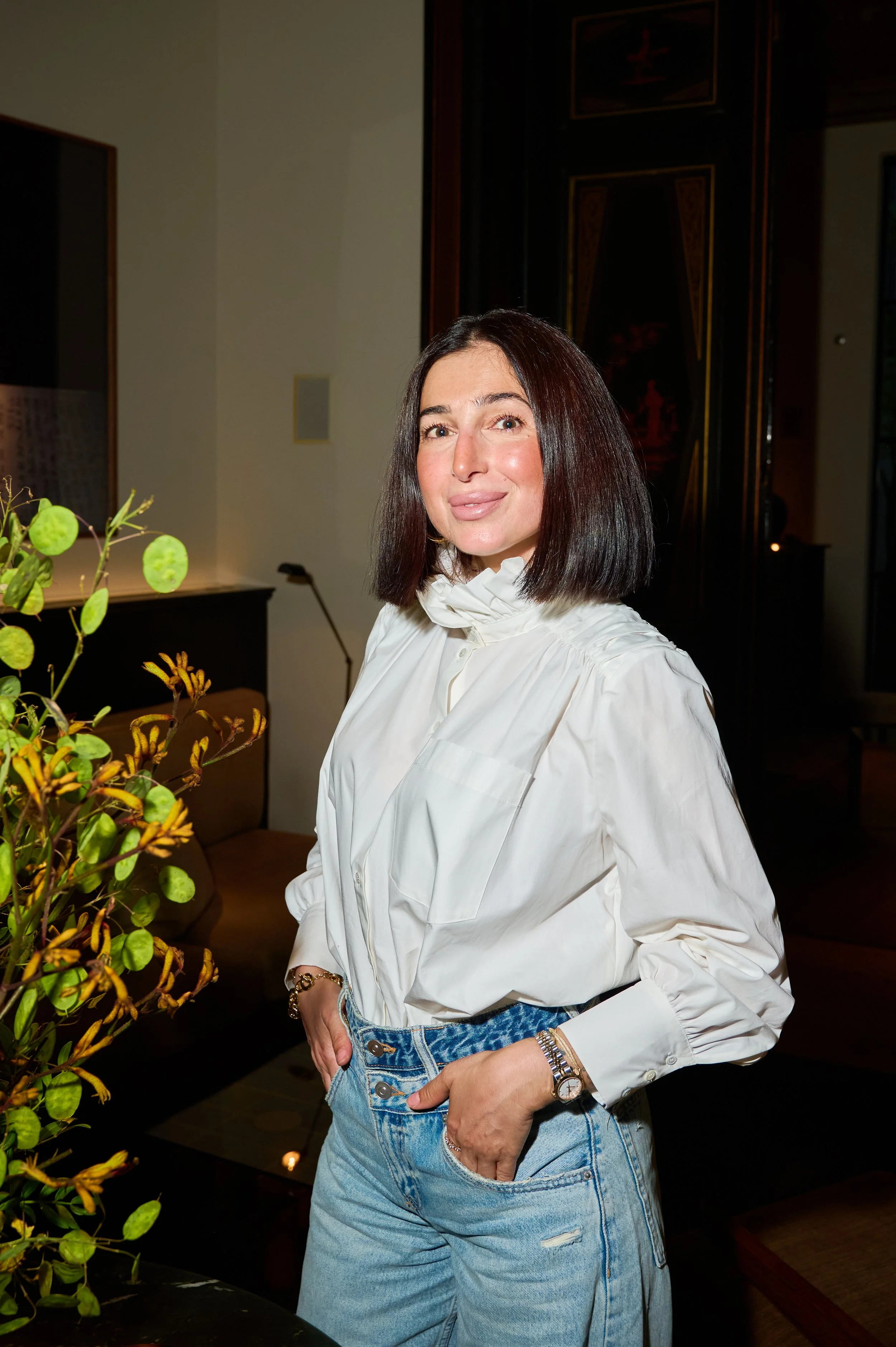 A woman with dark hair in a bob cut, wearing a white blouse and blue jeans, standing indoors with her hand in her pocket. There are plants in the foreground and dark furniture in the background.