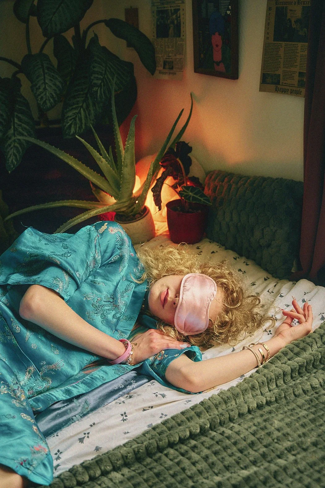 A woman with curly blonde hair is sleeping on a bed, wearing a pink sleep mask and a blue silk robe with floral embroidery. She has gold bracelets on her wrist and a pink bracelet on her other wrist. The bed has a white sheet with a small dark patter