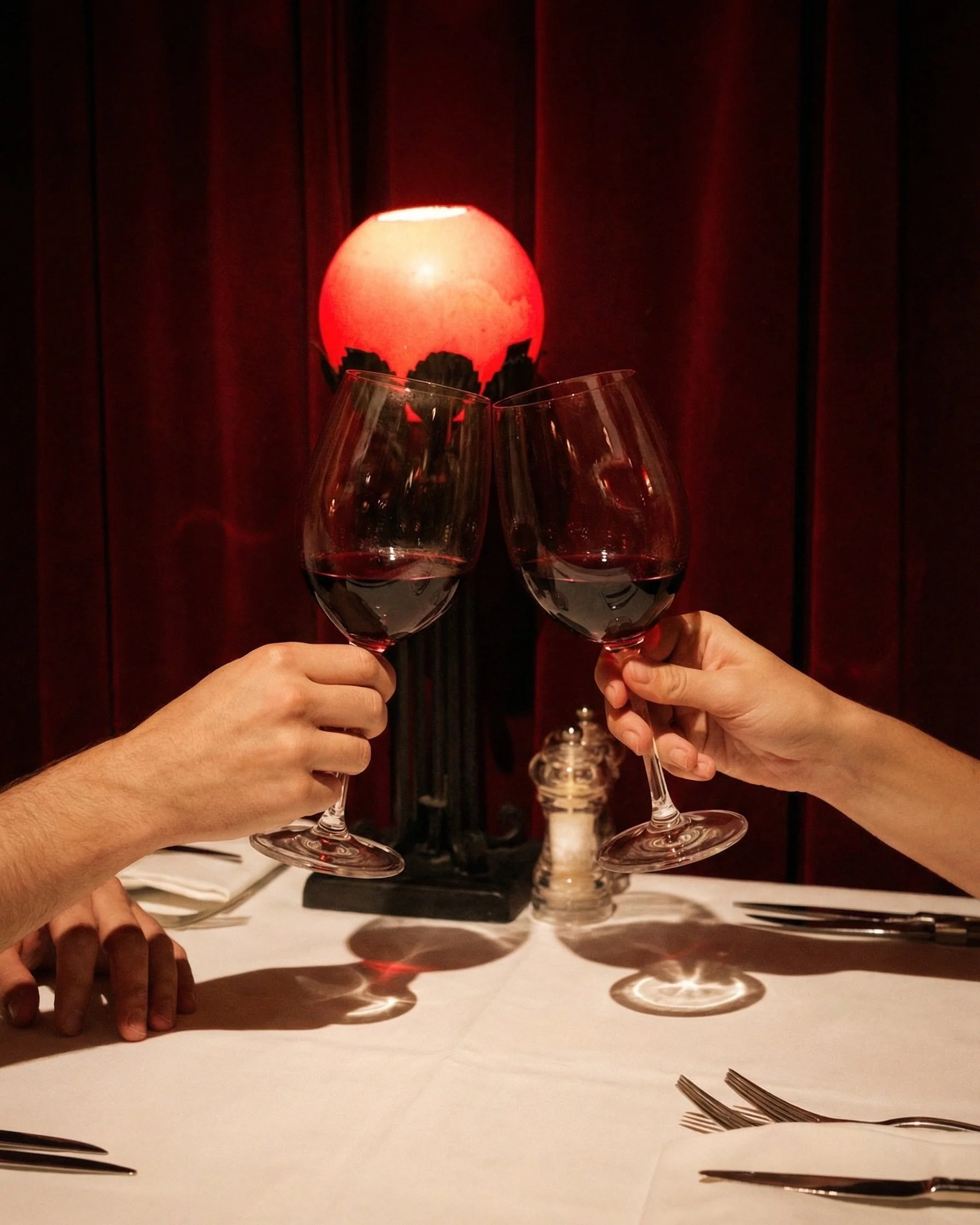 Two people clinking glasses of red wine at a dining table in a dimly lit restaurant with red curtains and a red lamp in the background.