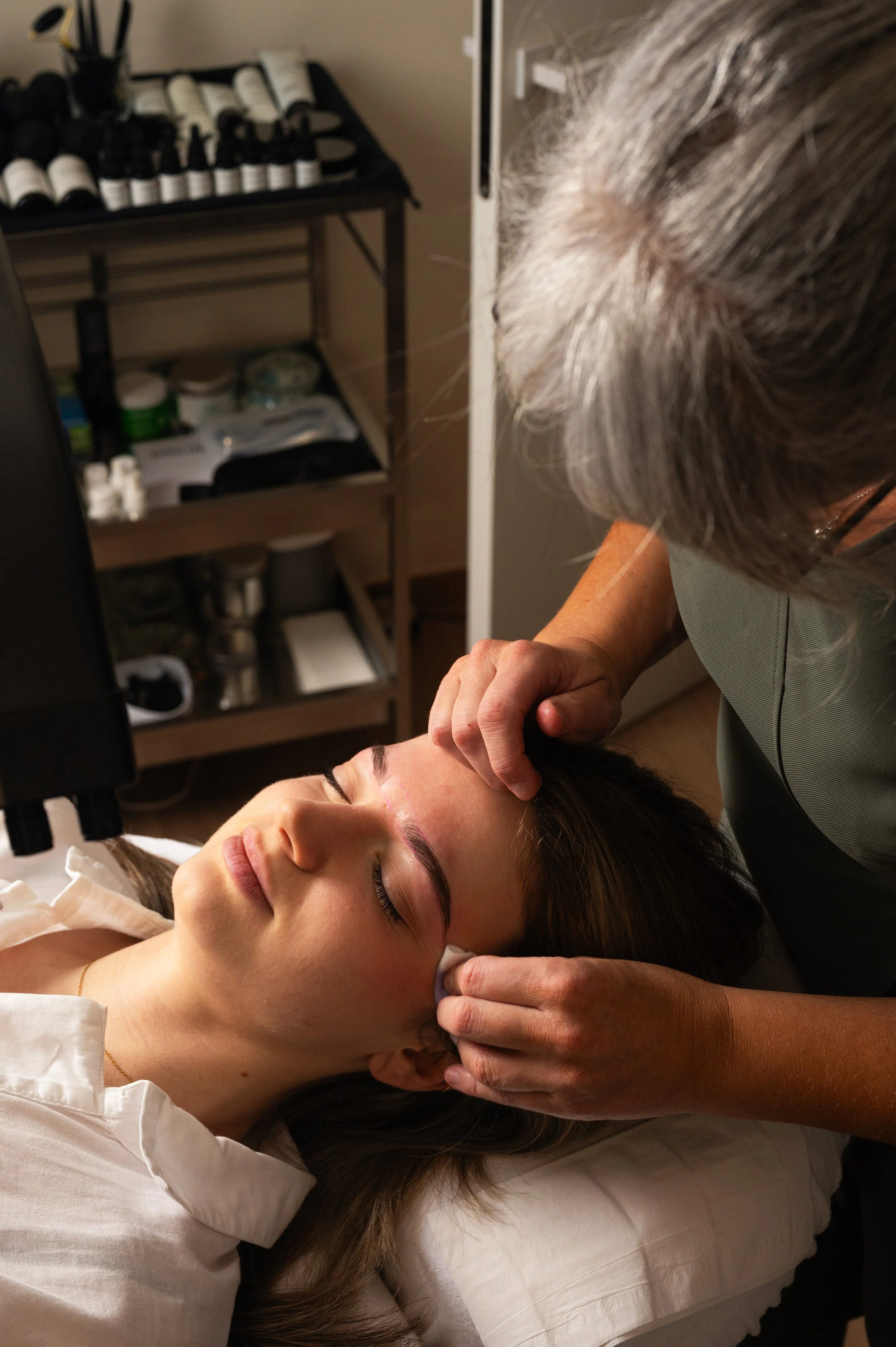 A woman lying on a medical bed with her eyes closed, receiving a skincare treatment or facial from a skincare professional who is gently applying a product to her forehead.