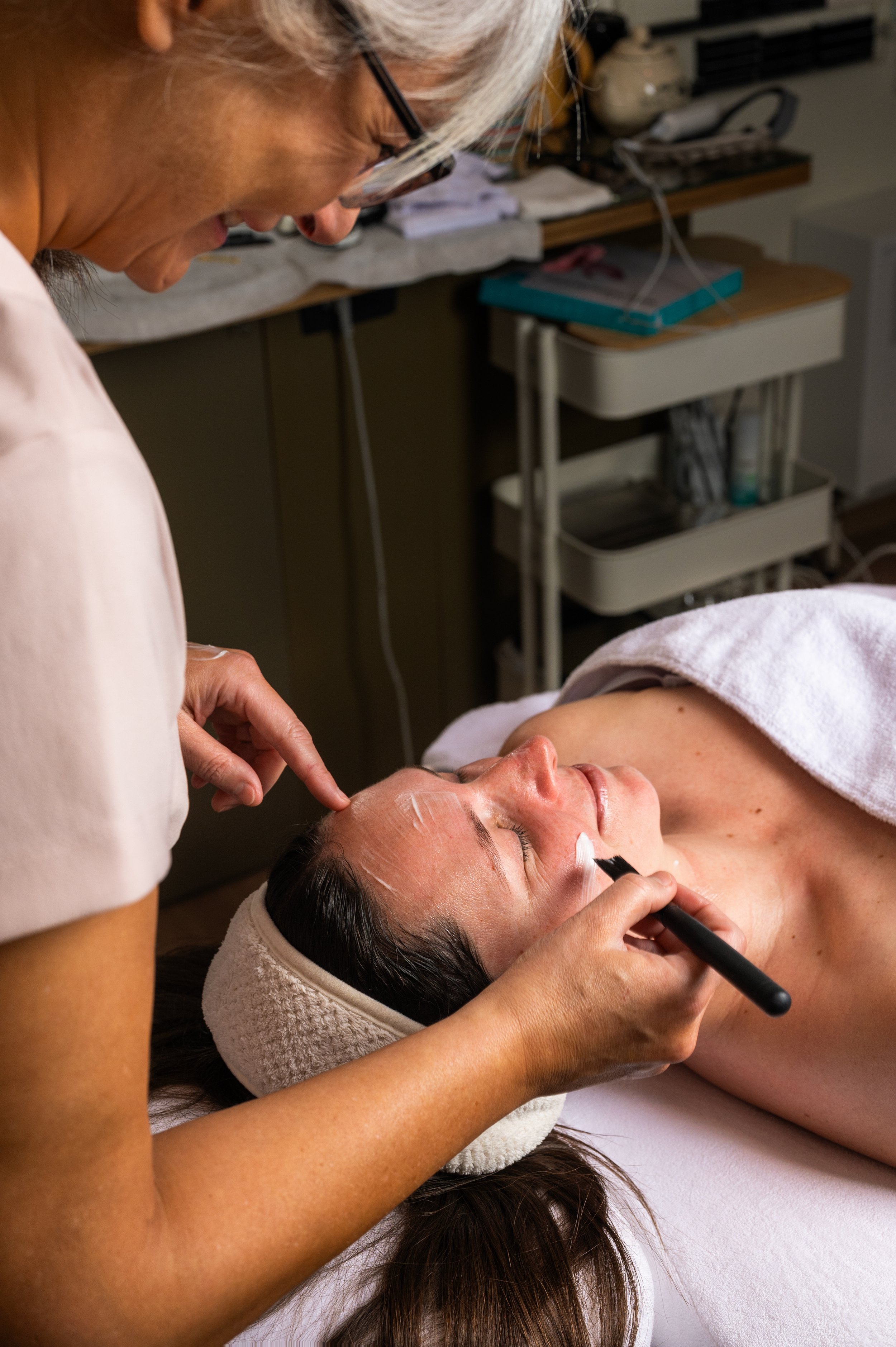 Esthetician applying a facial treatment to a woman lying on a spa bed, with a headband and a towel on her chest, in a spa or skincare clinic.