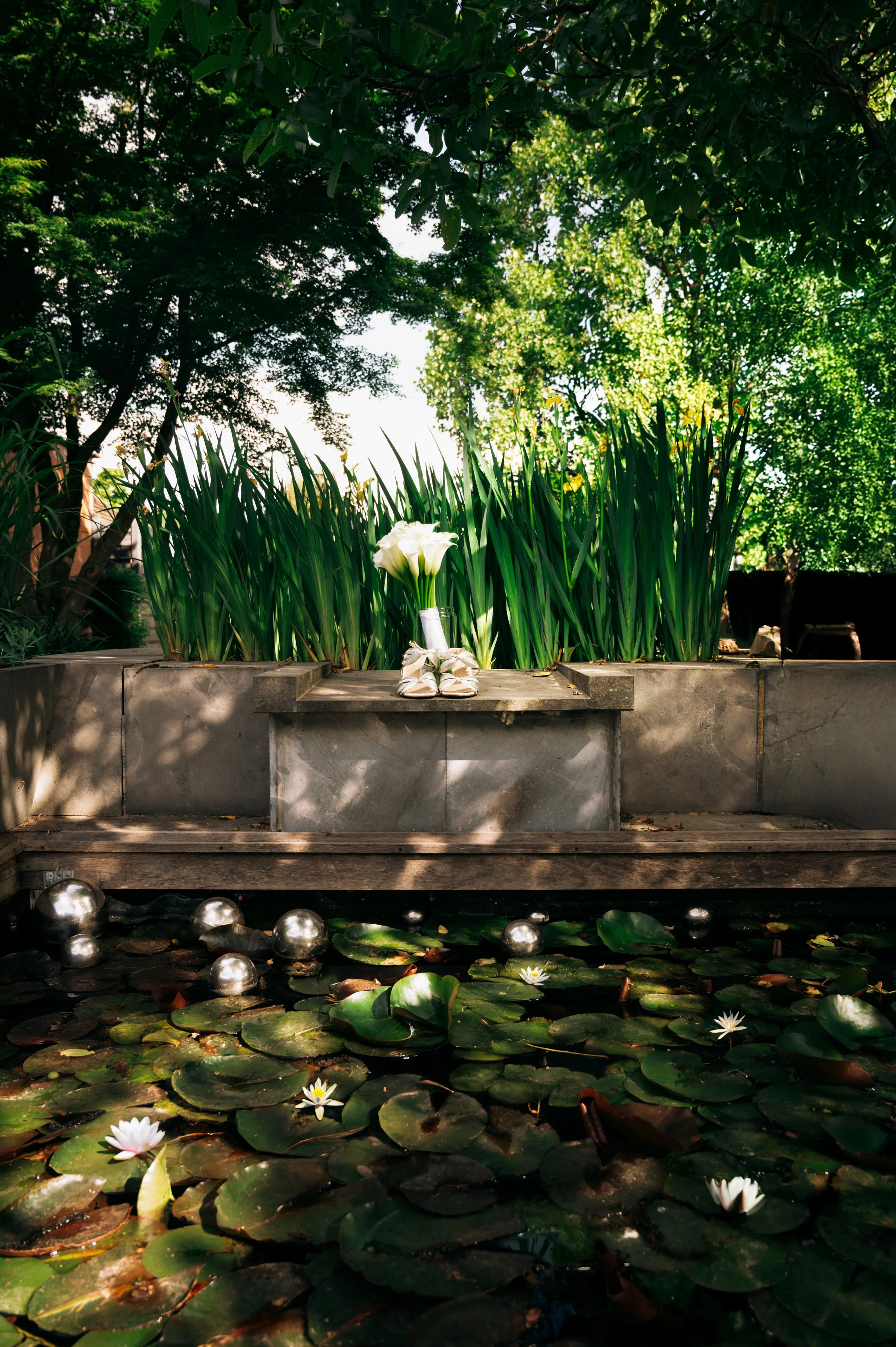 A serene outdoor scene with a small pond filled with lily pads and blooming white water lilies. In the background, a stone ledge holds tall green plants and a bouquet of white calla lilies, with a pair of shoes placed beside the bouquet. The scene is shaded by lush green trees with sunlight filtering through the leaves, creating a peaceful, natural atmosphere.