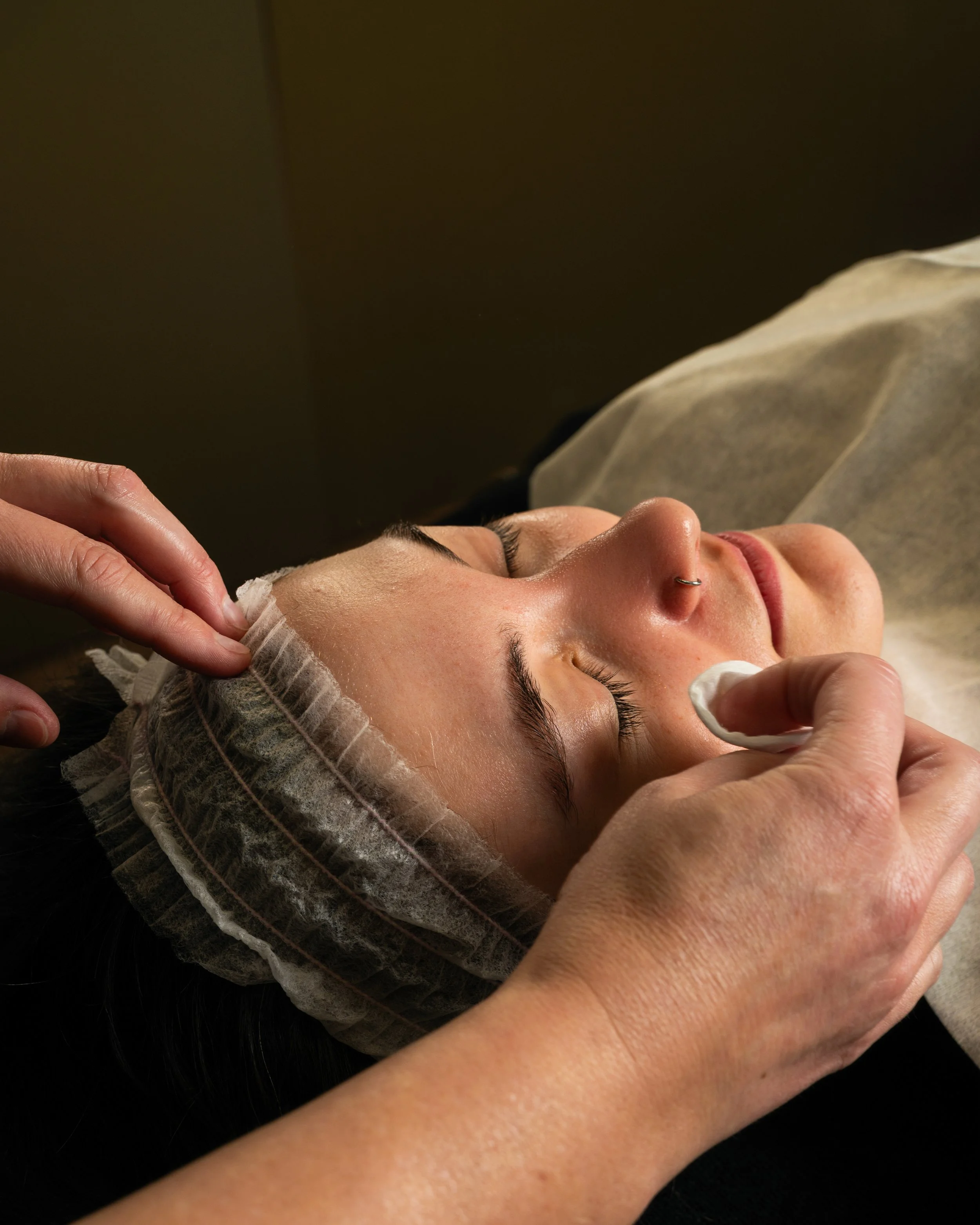Close-up of a woman with a nose piercing, receiving a facial treatment with a cotton pad while lying down, with someone gently applying it to her face.