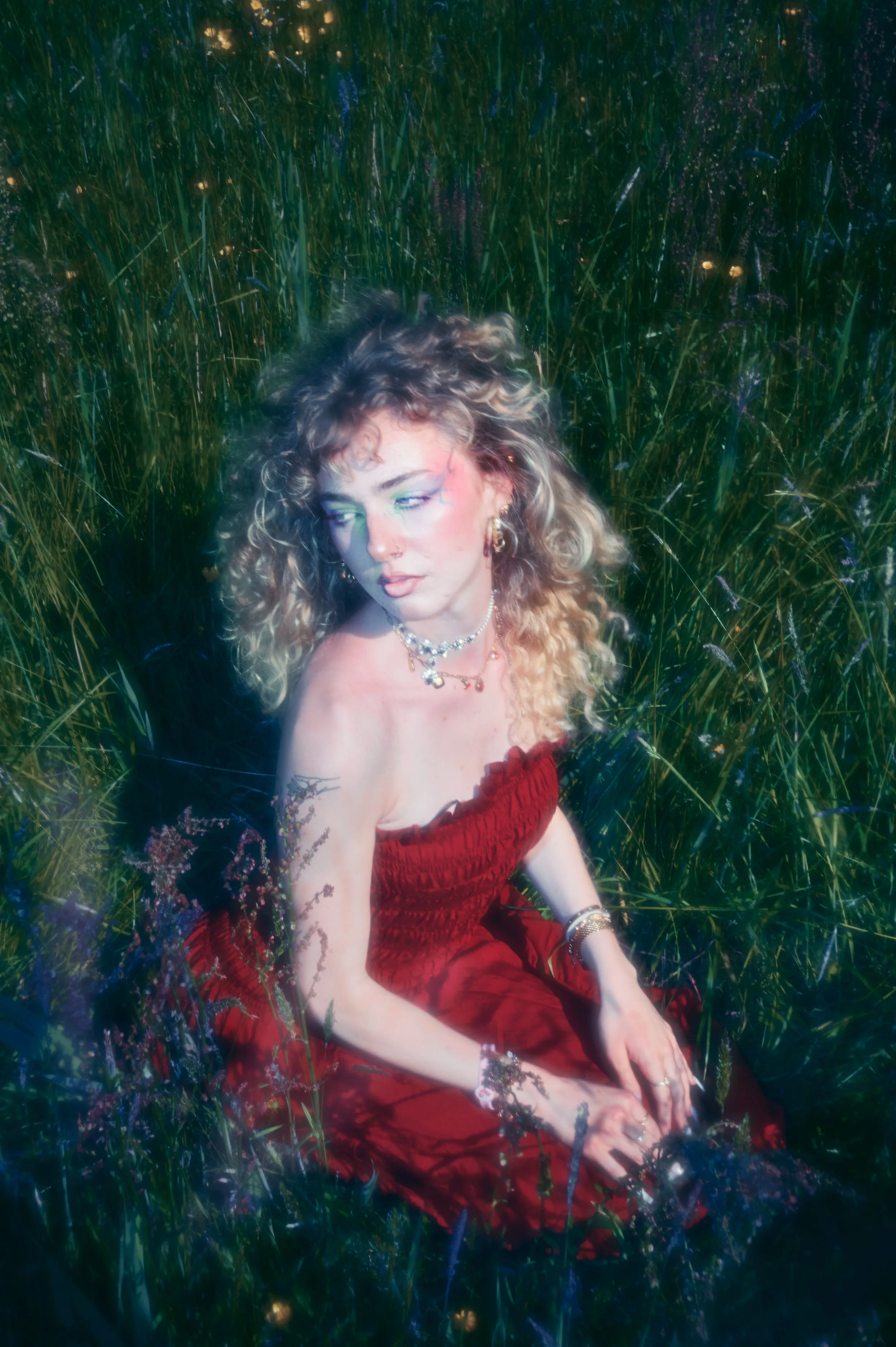 A woman with curly blonde hair in a red dress sitting in a field of tall grass and wildflowers, with a dreamy expression and jewelry, illuminated by soft light.