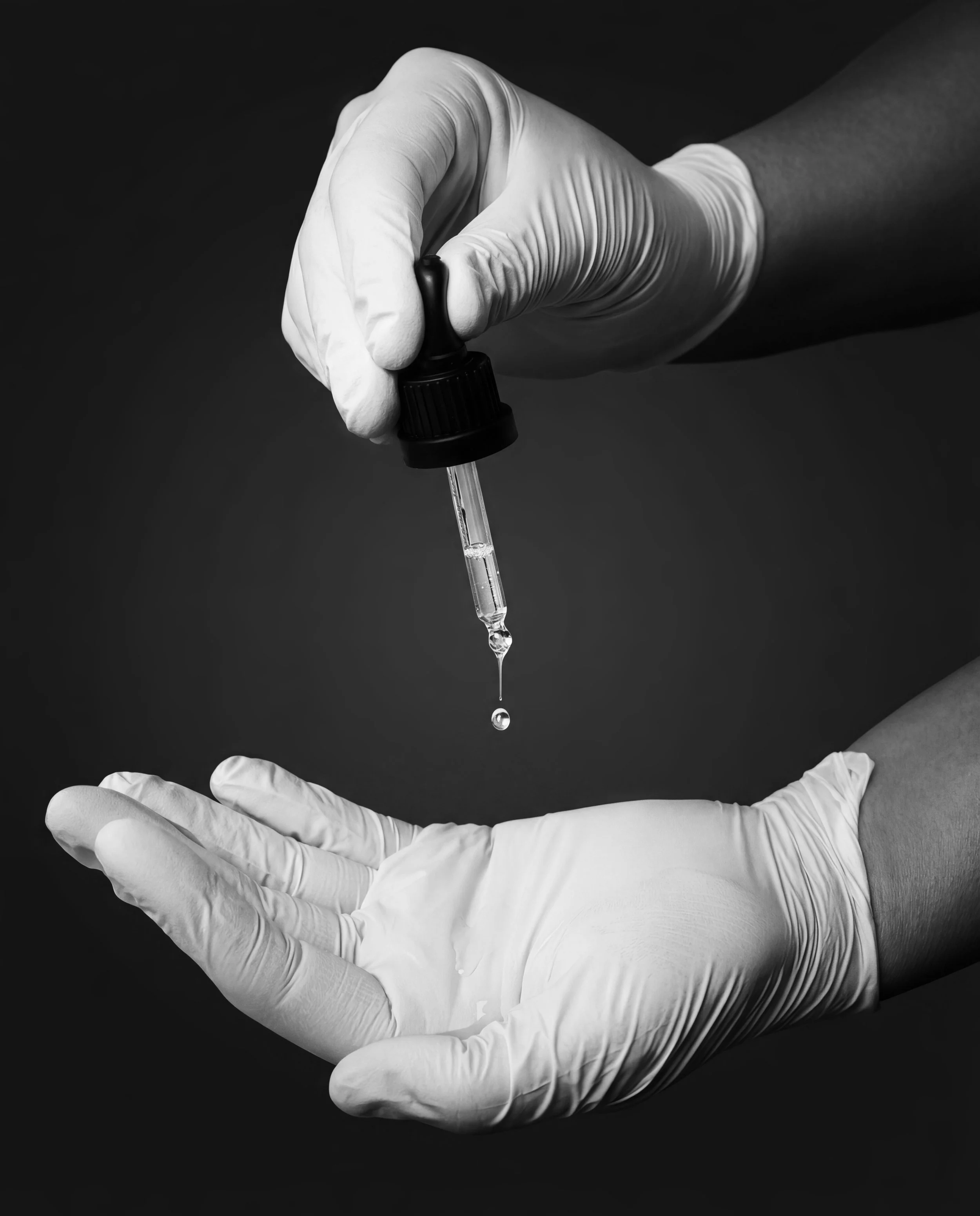 A person wearing white gloves holding a dropper with a liquid and a drop falling into an open palm.