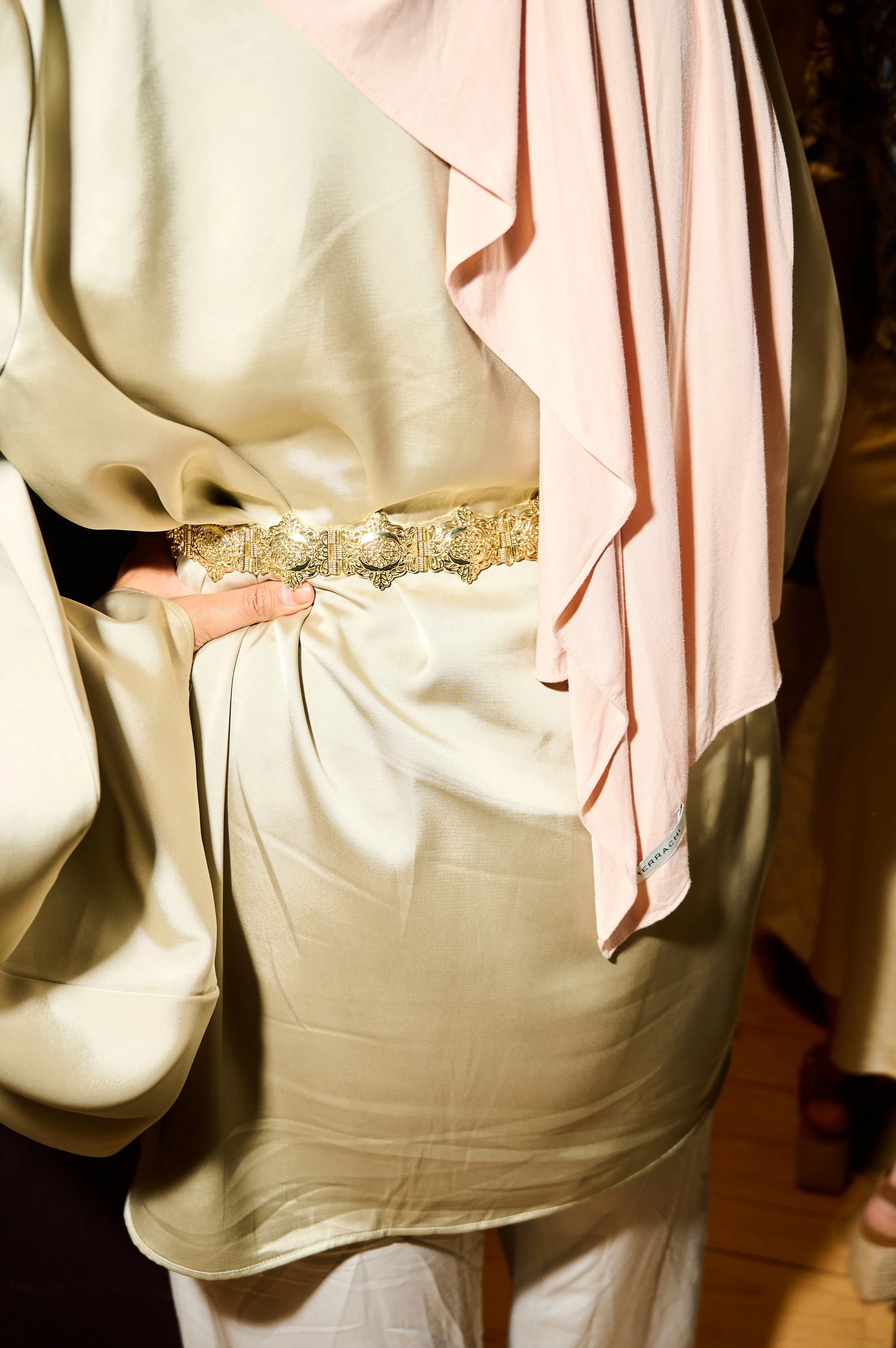 Close-up of a person wearing beige and light pink satin clothing with a gold embroidered belt, with a hand resting at the waist, and a pink satin shawl draped over the shoulder.