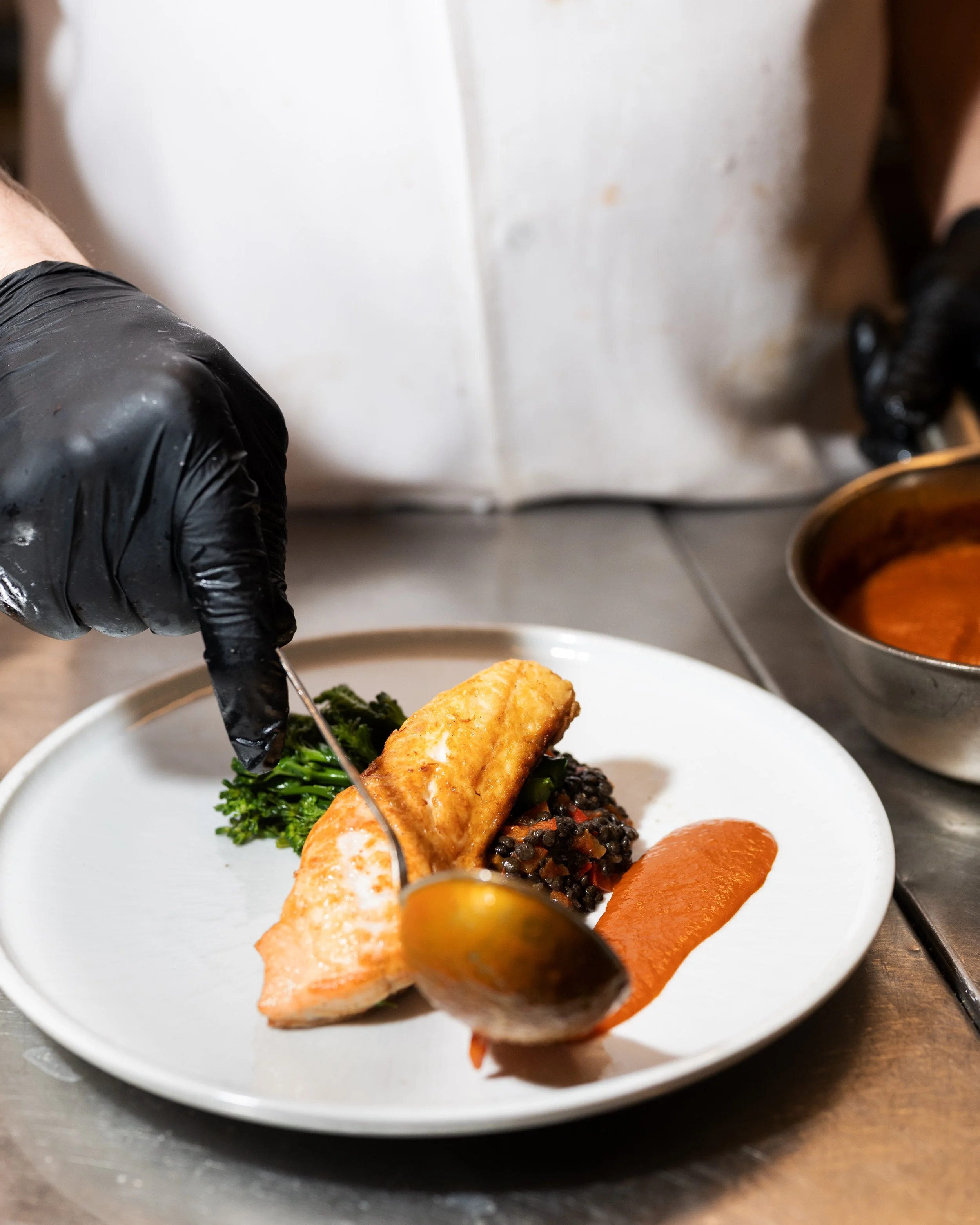 Chef garnishing a plated dish with a piece of cooked fish, served with black lentils, broccoli, and orange sauce.