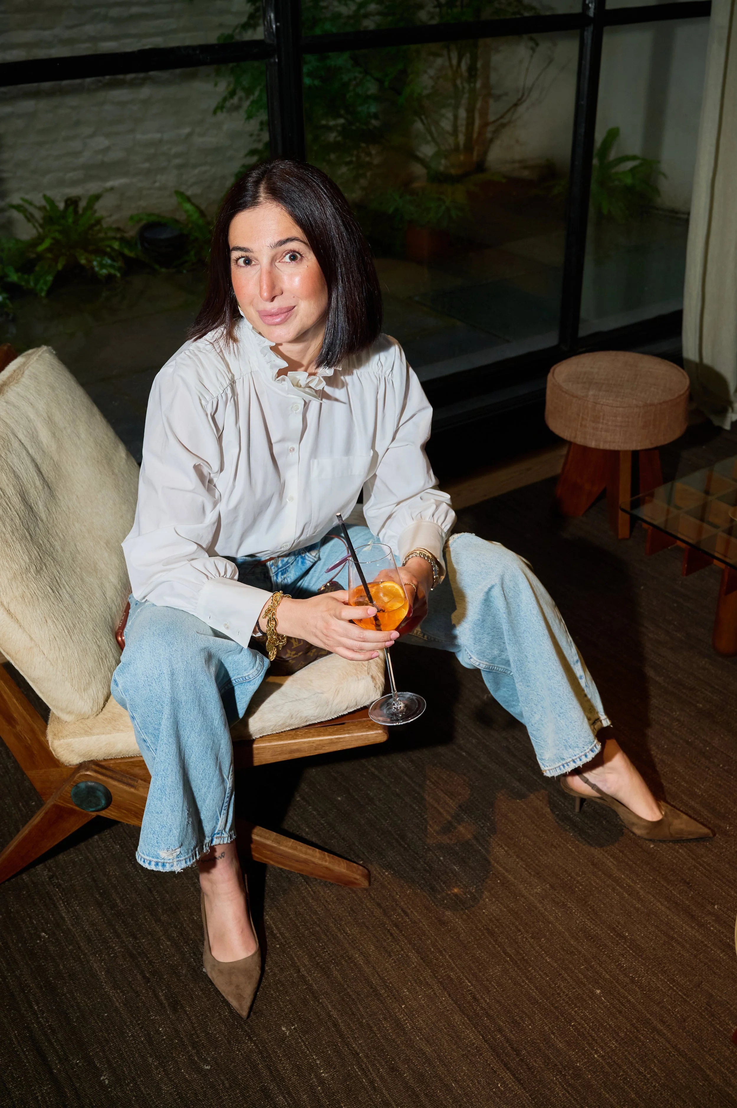 A woman with straight black hair sitting on a beige lounge chair in a dimly lit room. She is wearing a white blouse with ruffled collar, light blue jeans, and high heels. She is holding a glass of an orange-colored drink with a straw and smiling slig