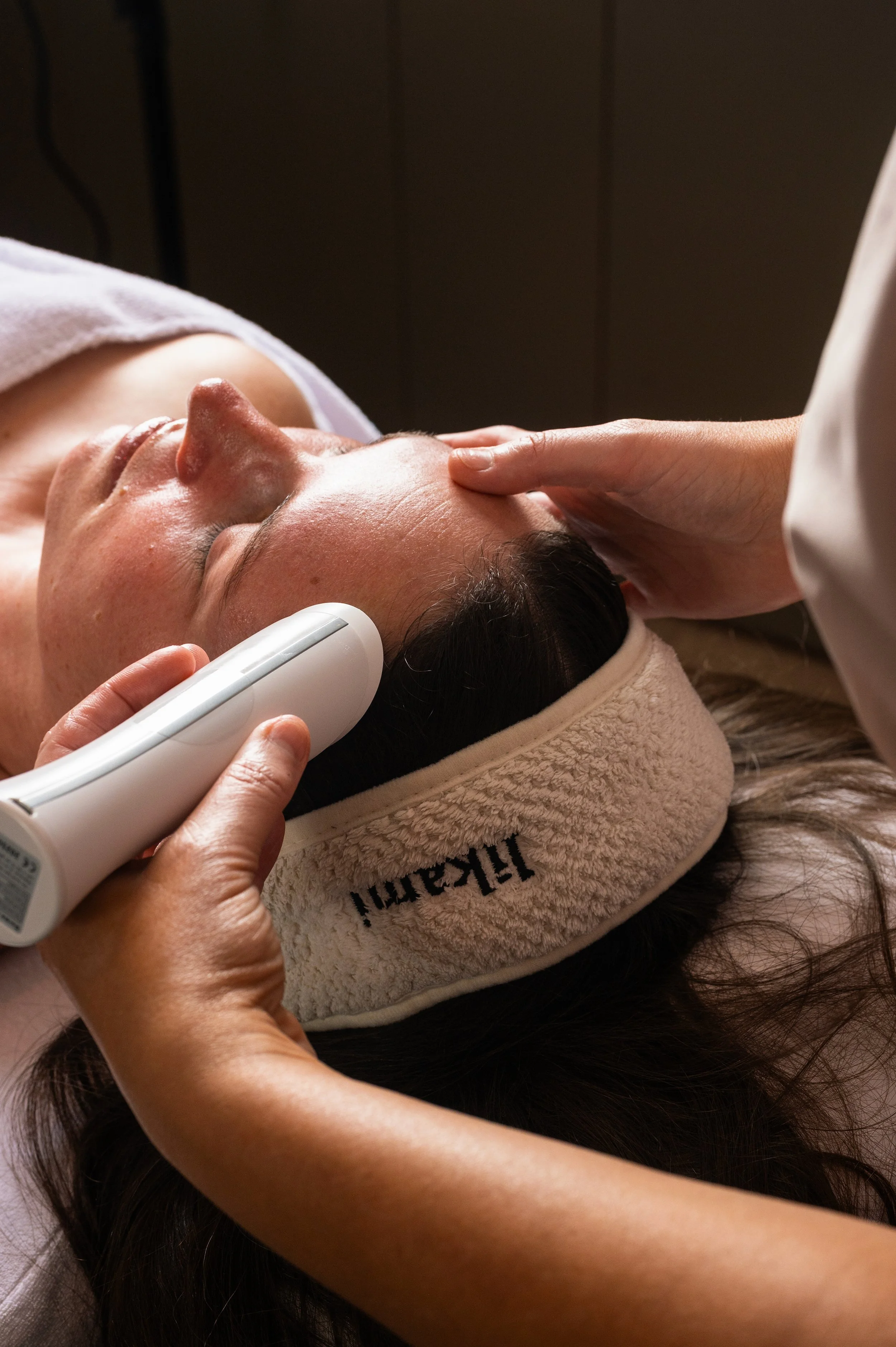A woman receiving a facial treatment with a handheld device while lying on a massage table, wearing a headband, in a spa setting.