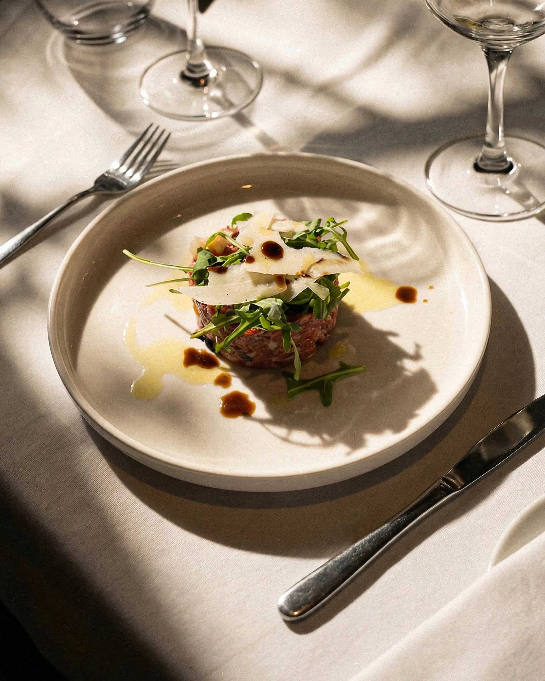 A close-up of a white plate with a small serving of food garnished with greens and shavings of cheese, drizzled with balsamic vinegar and olive oil. Silverware (fork and knife) on a white tablecloth, with empty wine glasses in the background.