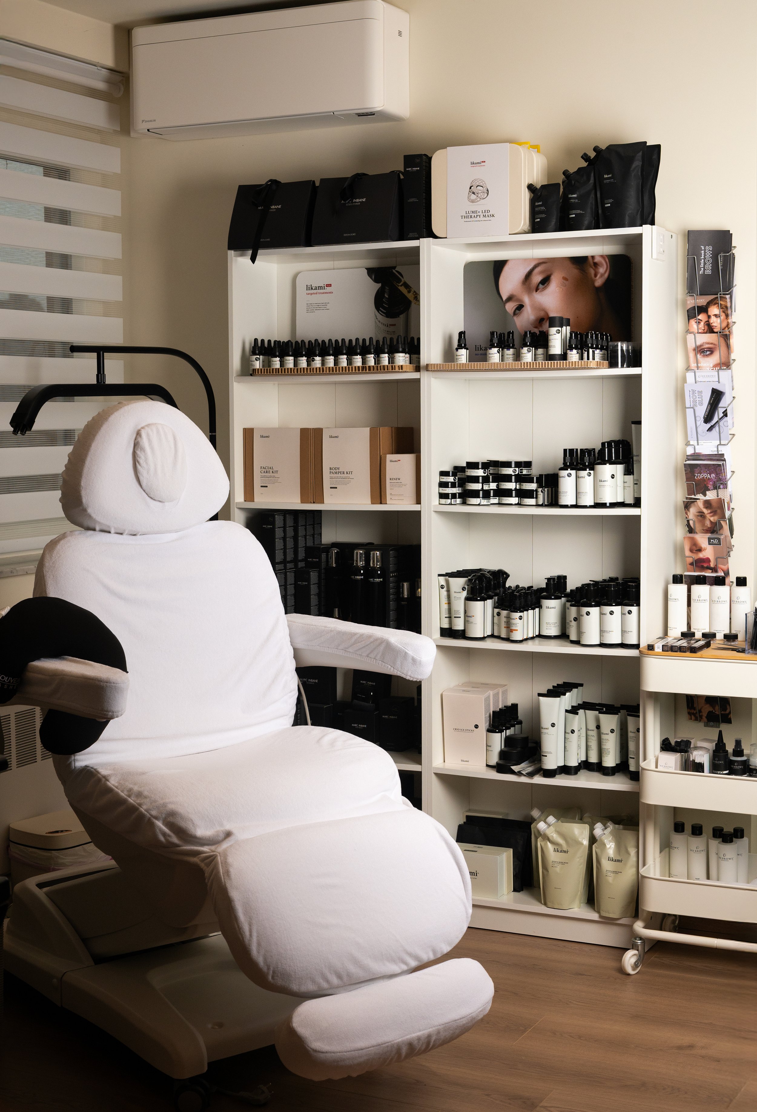 A treatment room with a white reclining chair wearing a headcover, facing a white shelf filled with skincare or beauty products, including bottles, jars, and boxes, in a spa or clinic setting.