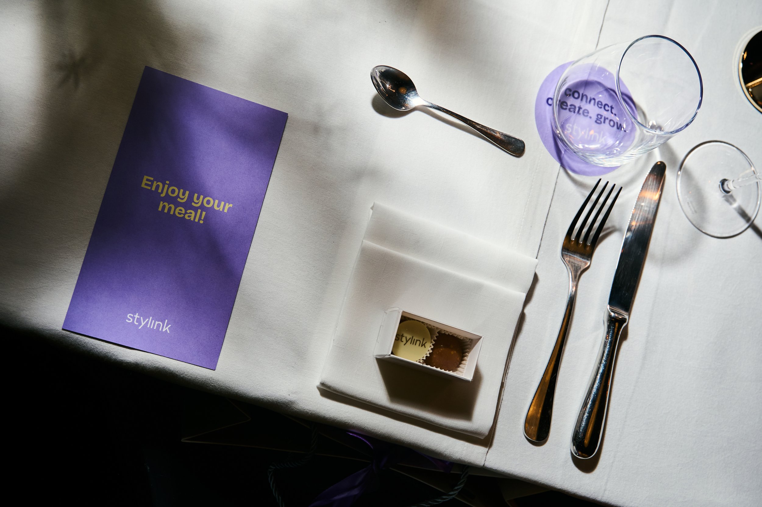 A white table setting with a purple napkin that says 'Enjoy your meal!', a small white box of chocolates with a 'stylink' logo, a water glass with purple drink and a 'connect. create. grow.' message, a silver spoon, fork, and knife, and a napkin.